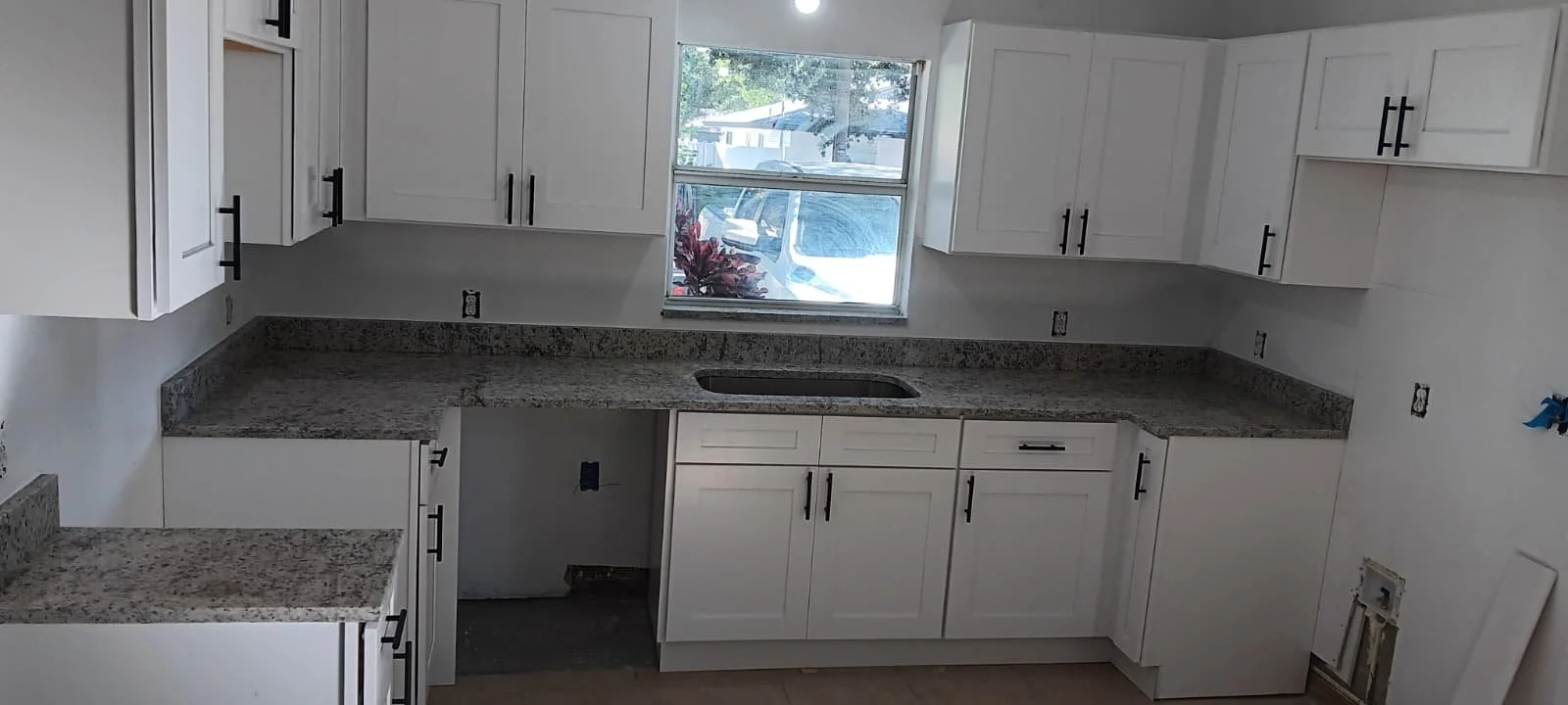 Kitchen with white cabinets, granite countertops, and a window above the sink. The cabinet hardware is black, and there are missing appliances and outlets.