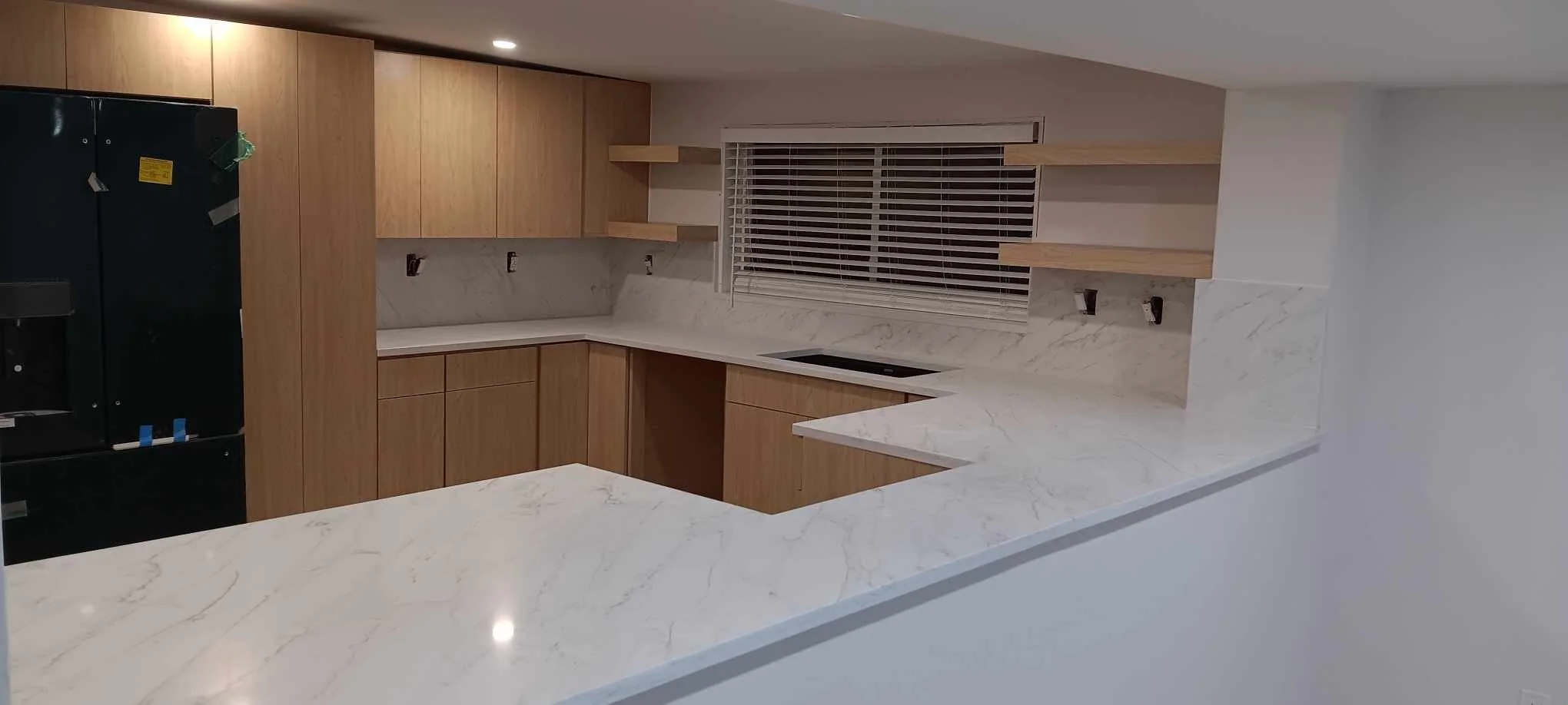 Modern kitchen with wooden cabinets, marble countertops, and a window with blinds. Open shelving above the counter and a black refrigerator on the left.
