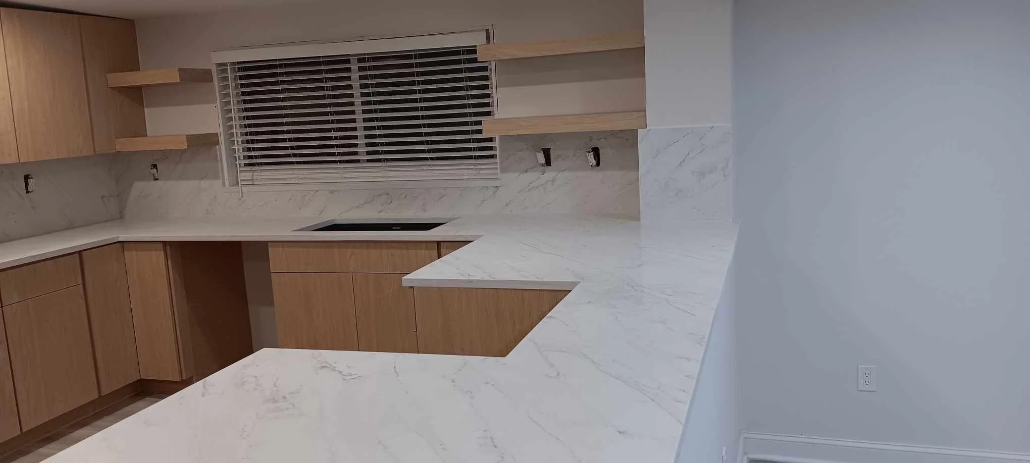 Empty kitchen with light wood cabinets, white marble countertops, a black cooktop, and a window with blinds. Shelves are mounted above the window, and the wall is painted white.