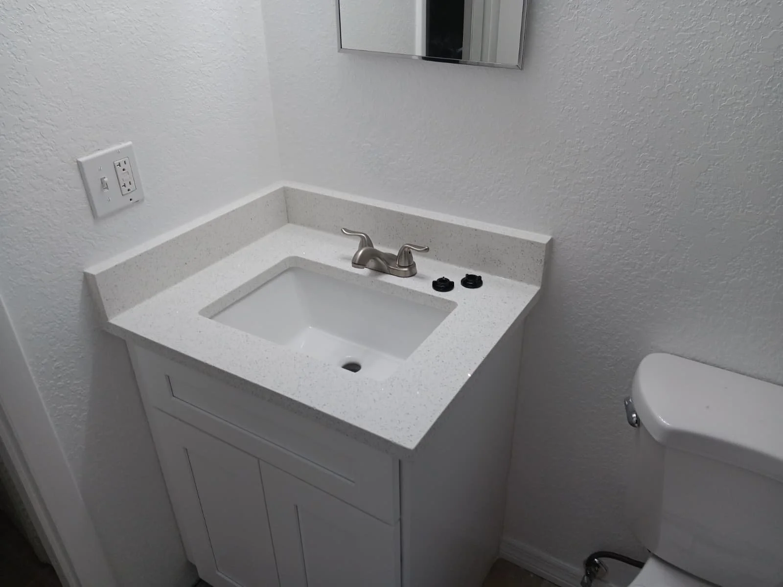 Small white bathroom sink with a silver faucet, black handles, and a white cabinet underneath, next to a white toilet, in a plain white-tiled room.