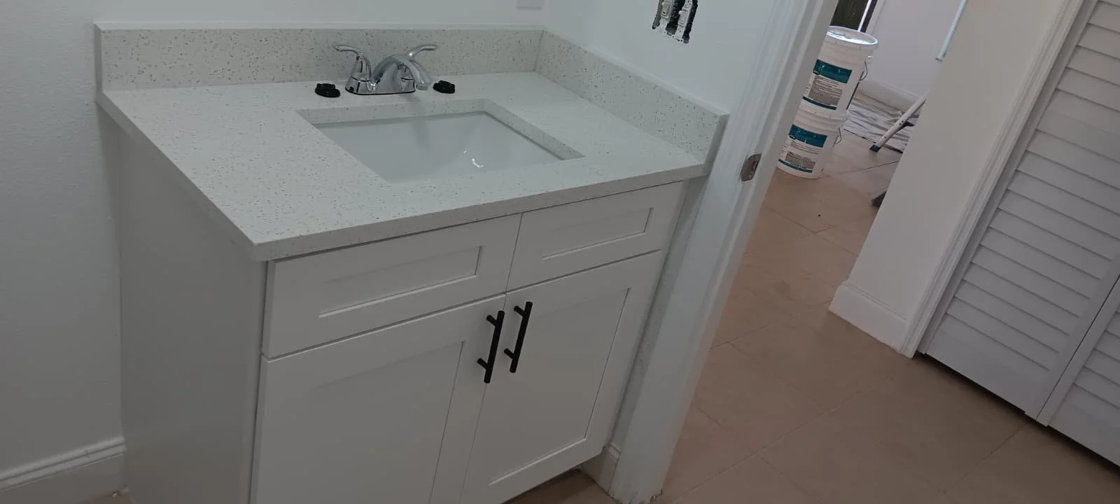 White bathroom vanity with a sink, faucet, black handles, and a speckled white countertop, with a doorway leading to the laundry area in the background.