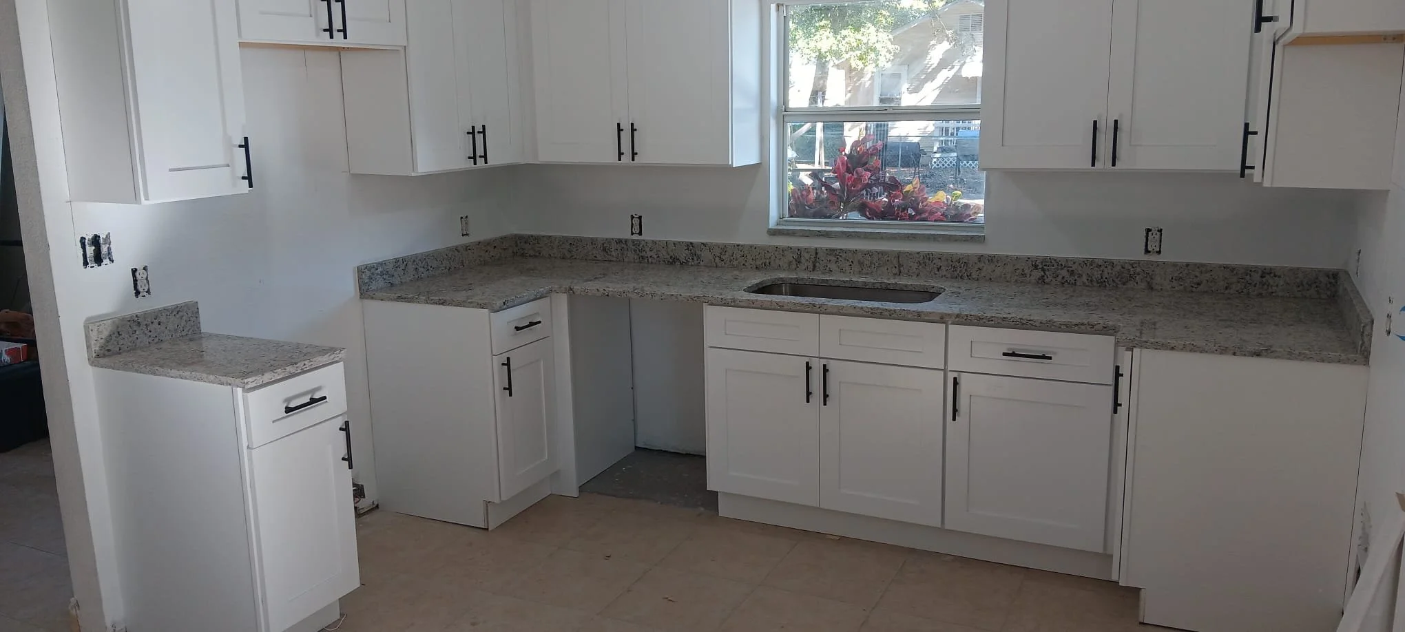 Kitchen with white cabinets, granite countertops, and a window above the sink showing a tree and a house outside, in the process of renovation.
