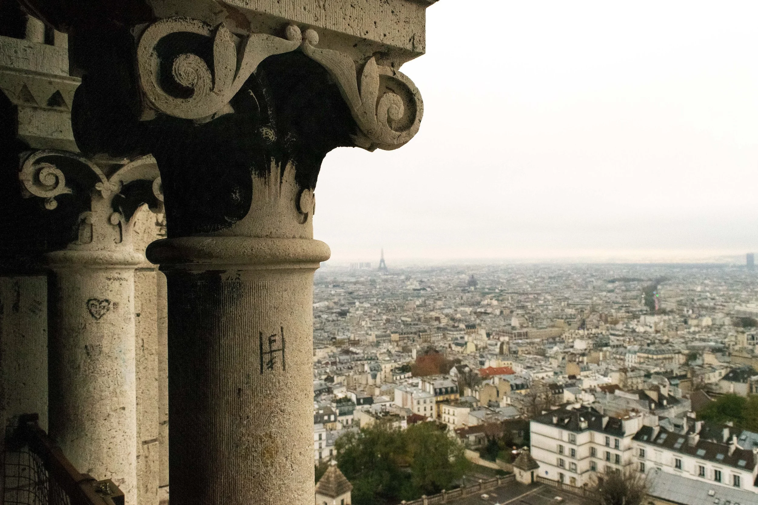 Urban scene with an industrial brick building and smokestack, steel bridge structure on the left, cars on the street, and modern high-rise buildings in the background under a partly cloudy sky.