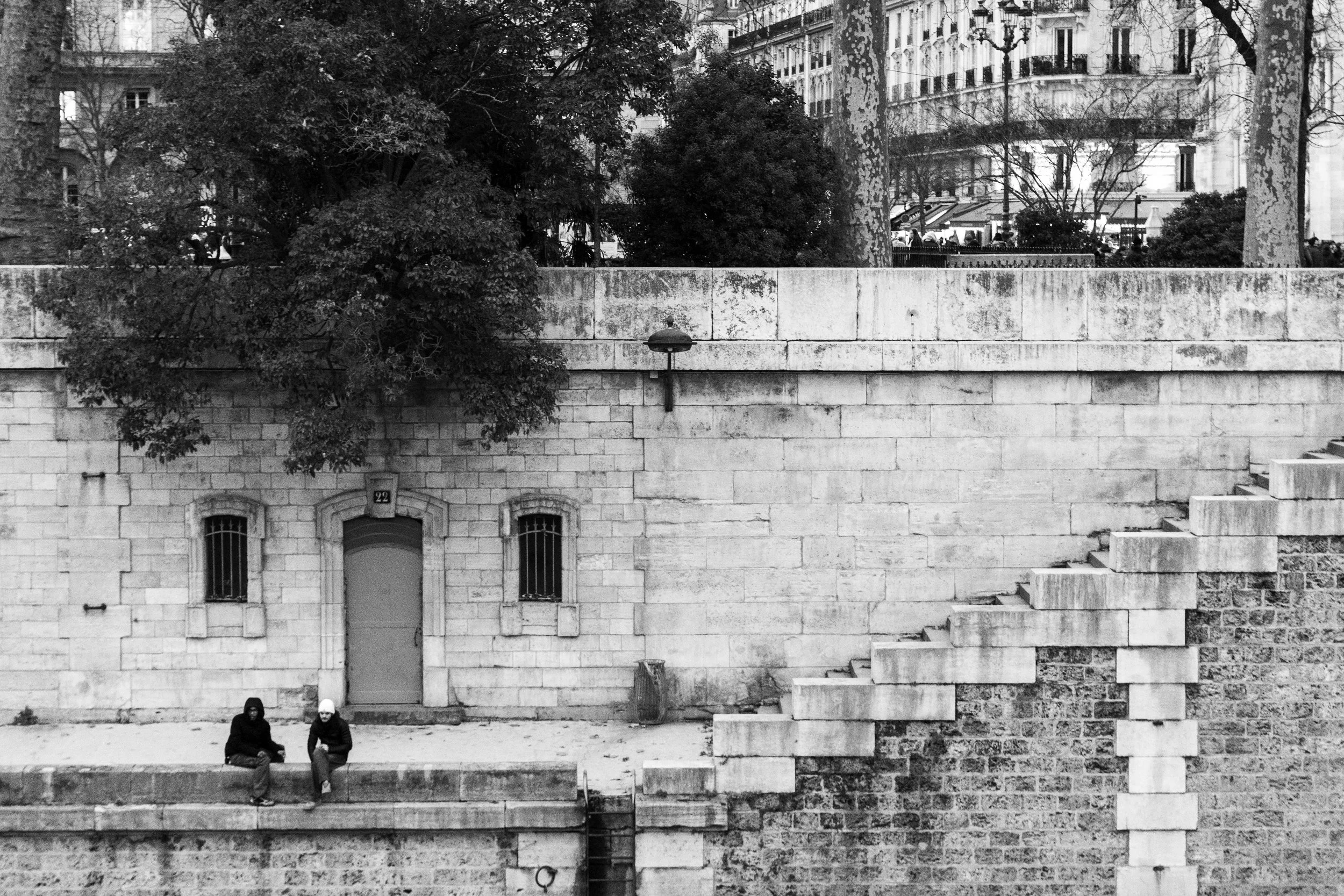 Black and white photo of a busy city street corner with people walking and sitting, tall buildings, and the Flatiron Building in the background at the intersection of Broadway and 5th Avenue.