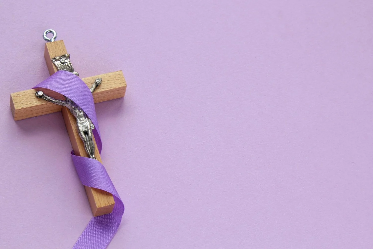 Wooden cross with a small crucifix attached, wrapped with a purple ribbon, placed on a lavender background.