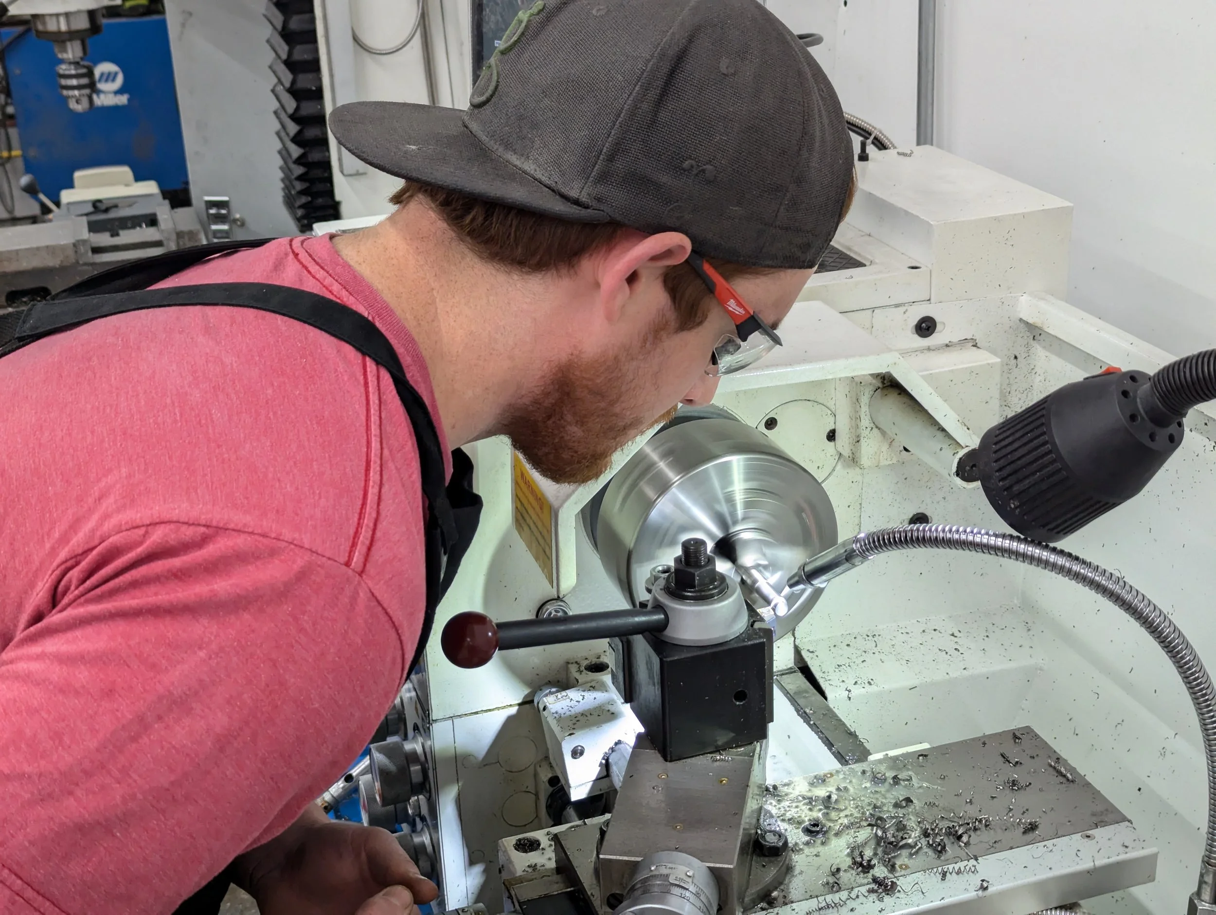 A man wearing safety glasses and a gray cap operates a metal lathe, shaping a piece of metal. The lathe is white and has metal shavings on the tray below the workpiece.