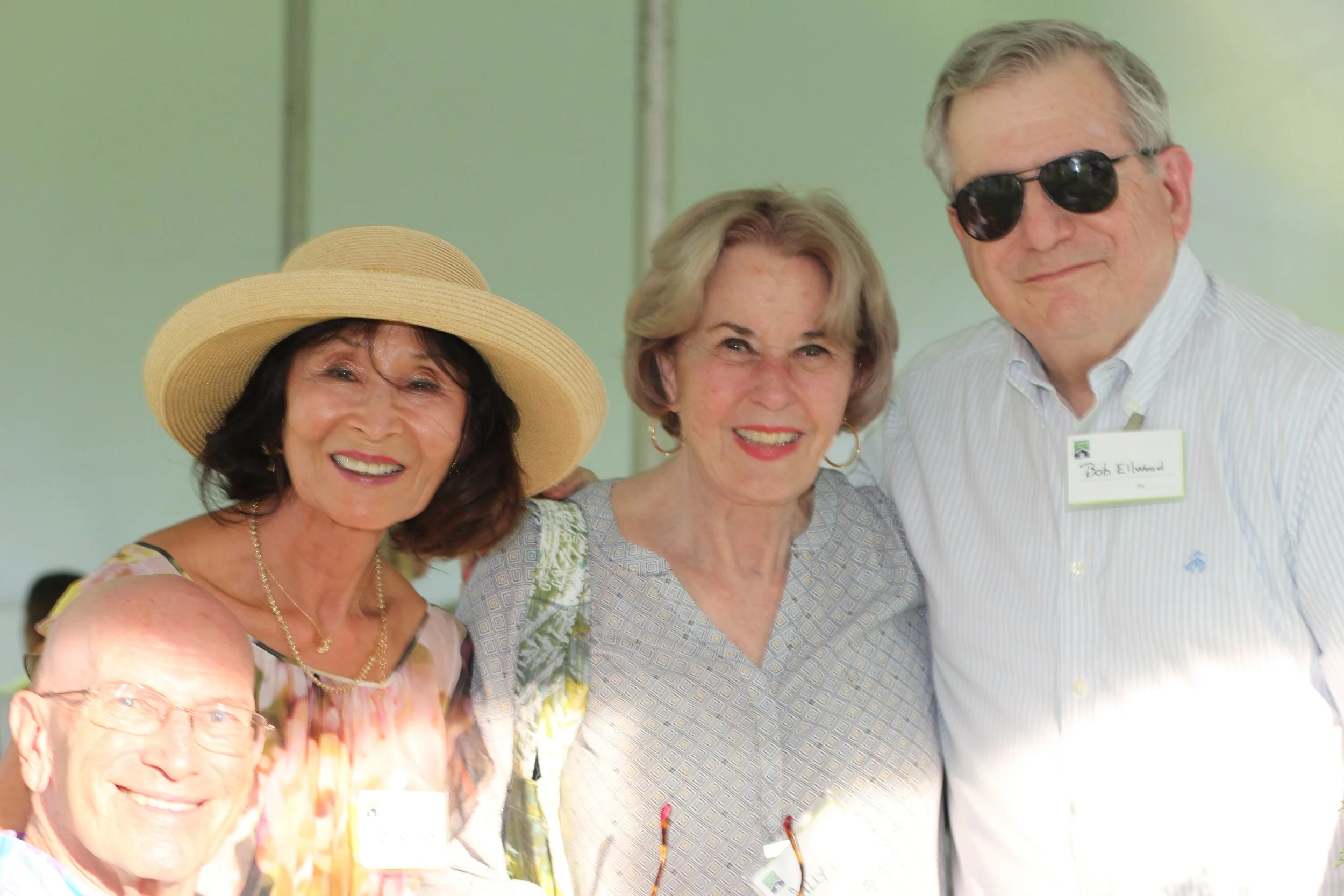 Four smiling older adults at a social gathering in Litchfield County for Chore Service, two women and two men, with one woman wearing a large sun hat and the man wearing sunglasses, standing close together.