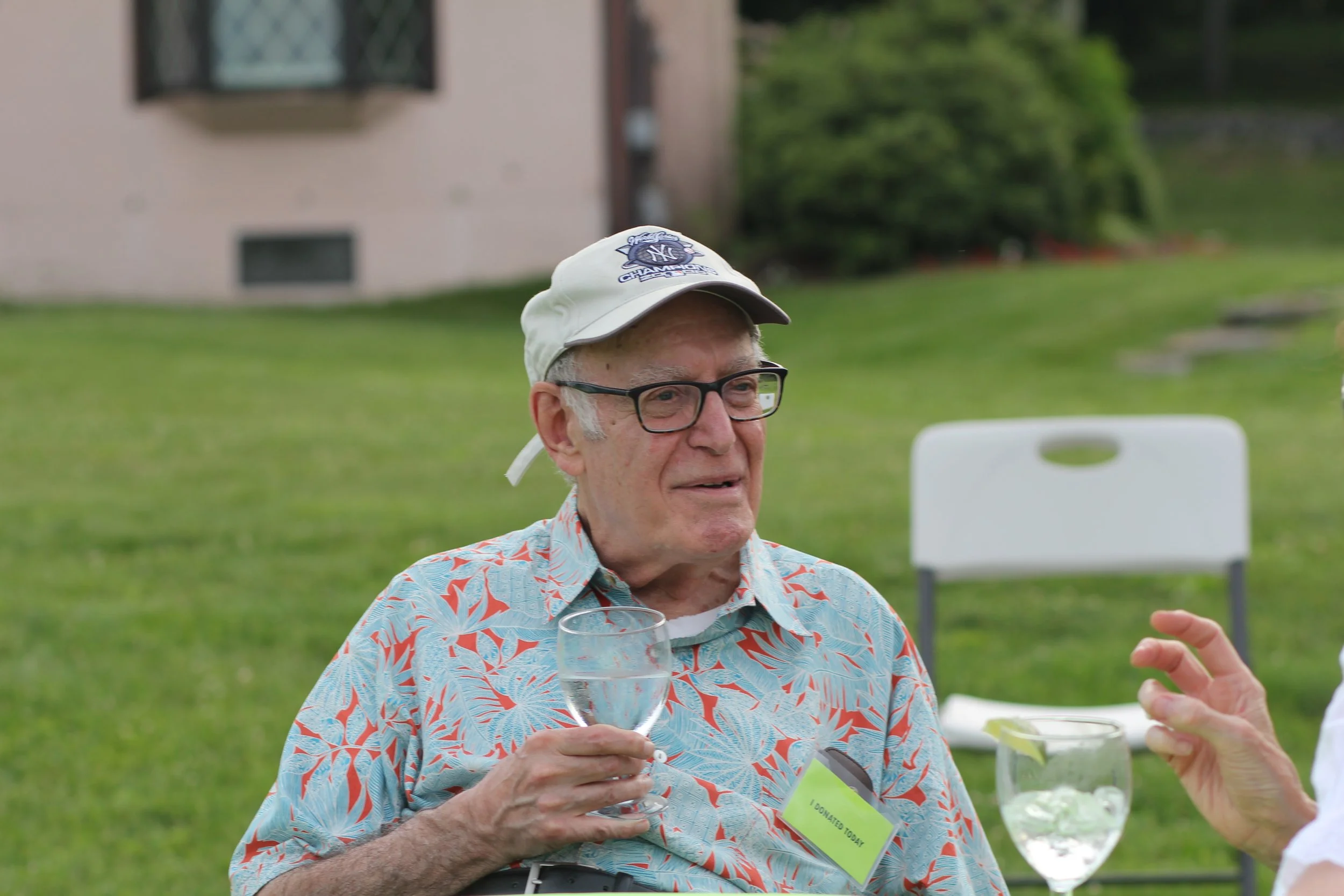 An elderly man with glasses, wearing a baseball cap and a colorful Hawaiian shirt, holding a glass of water, outdoors at a gathering in Litchfield County. A white chair is behind him and another person’s hand is visible gesturing.