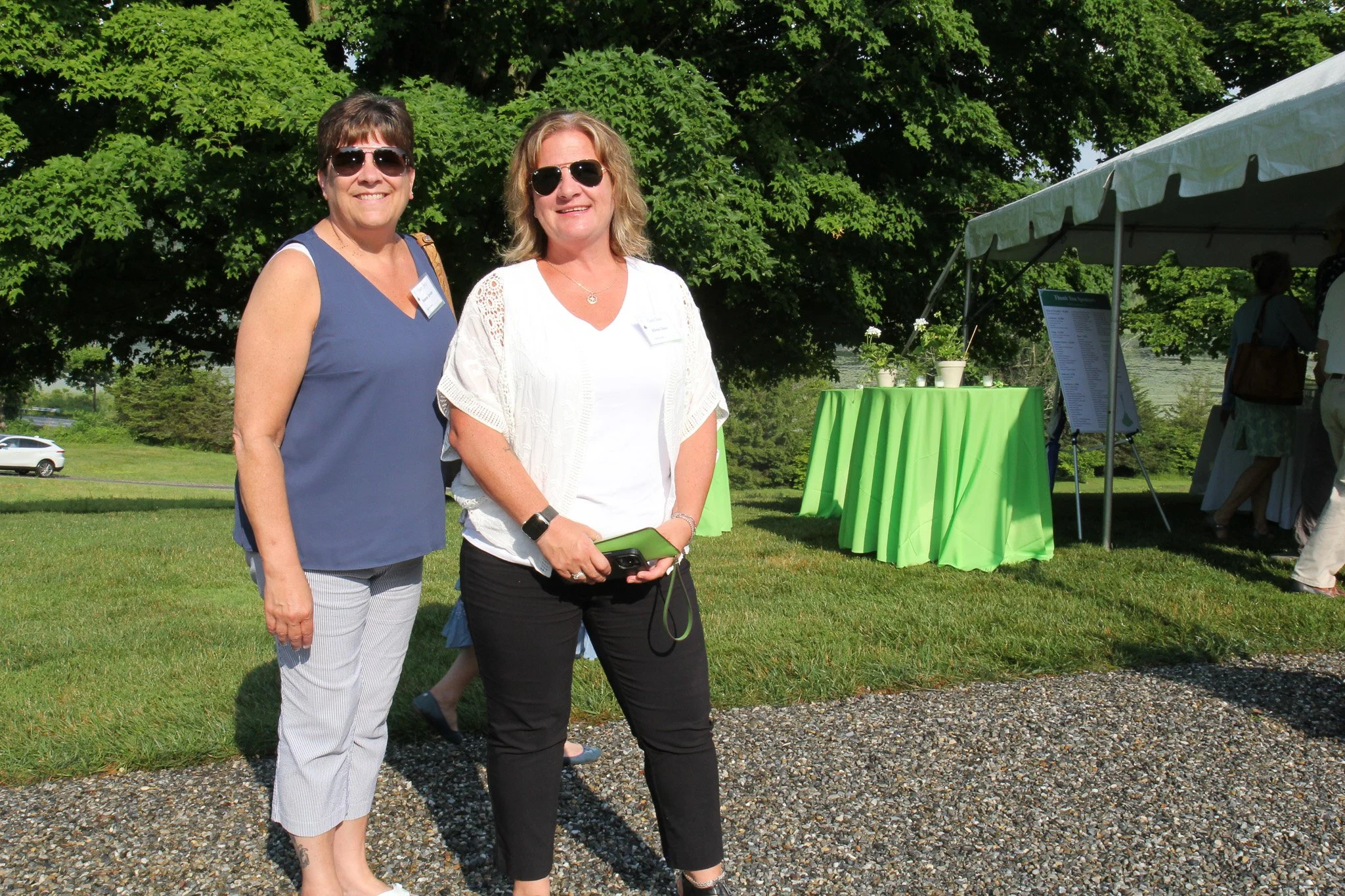 Two women standing outdoors on grass near a tent with a green tablecloth and potted plants, smiling at the camera, wearing sunglasses.
