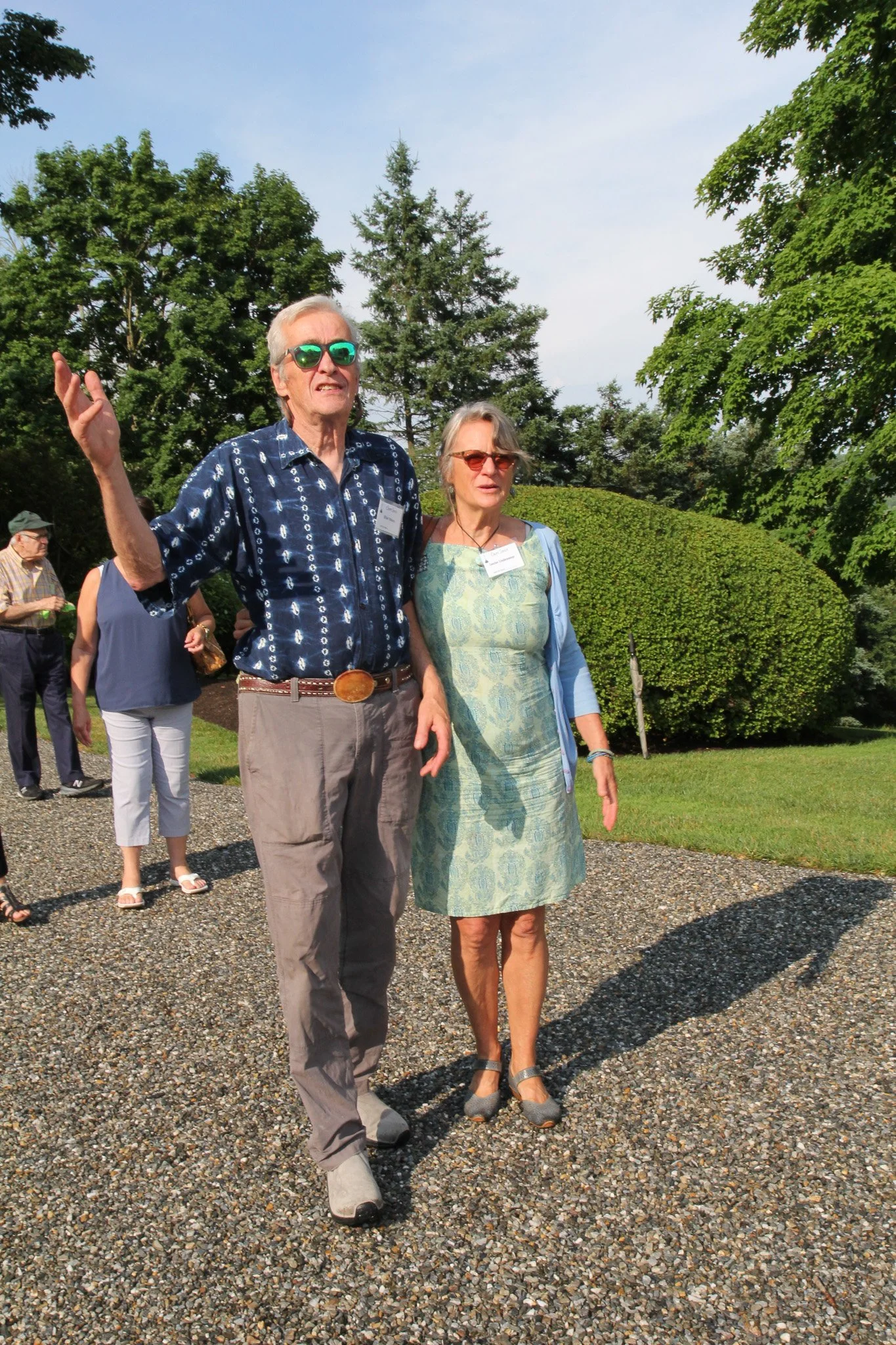 Two elderly people walking outdoors on a gravel path with other people in the background, surrounded by green trees and bushes, under a partly cloudy sky.