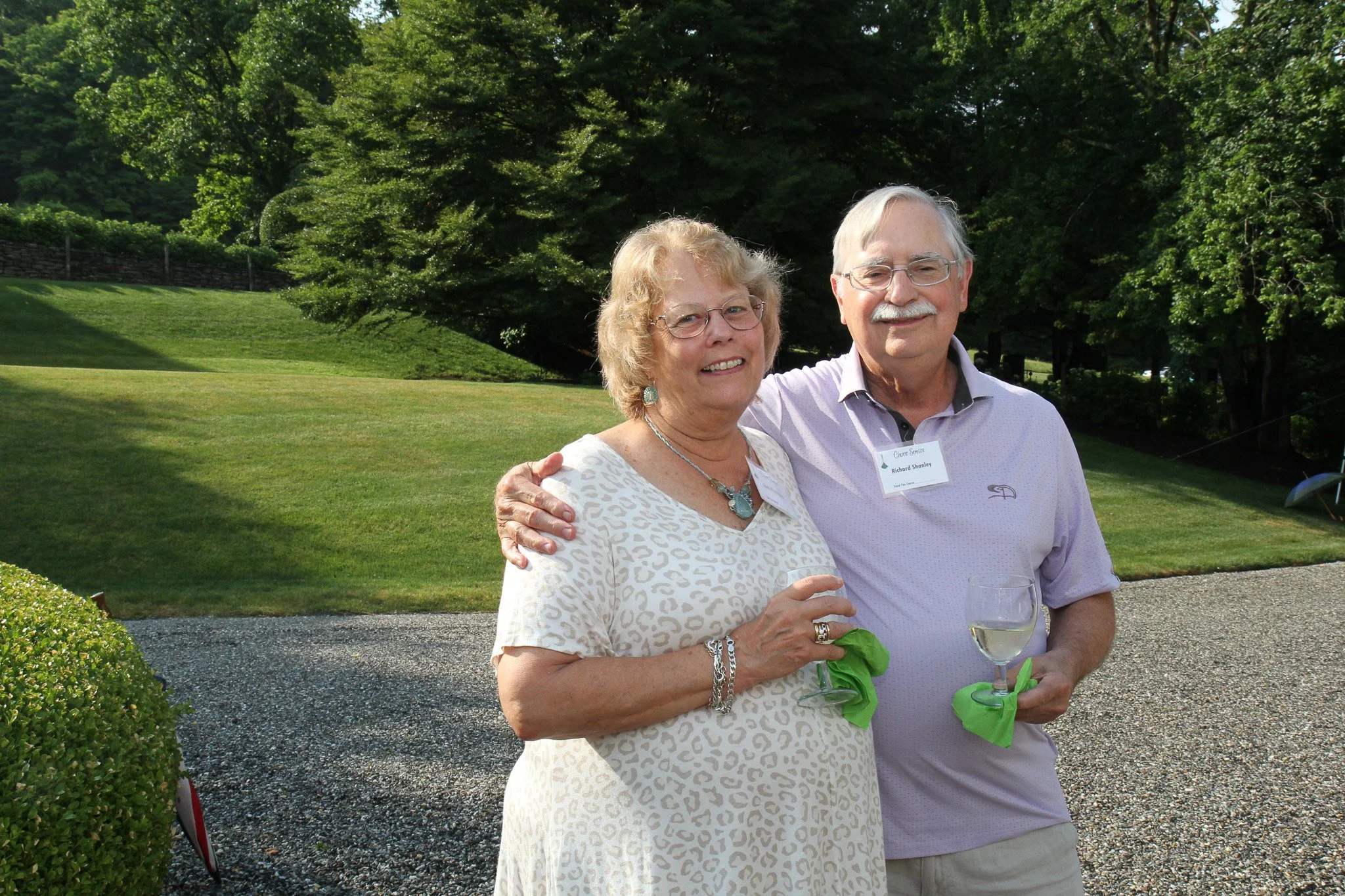 An elderly woman and man smiling outdoors at a garden party, holding glasses of white wine, with a lush green landscape in the background.