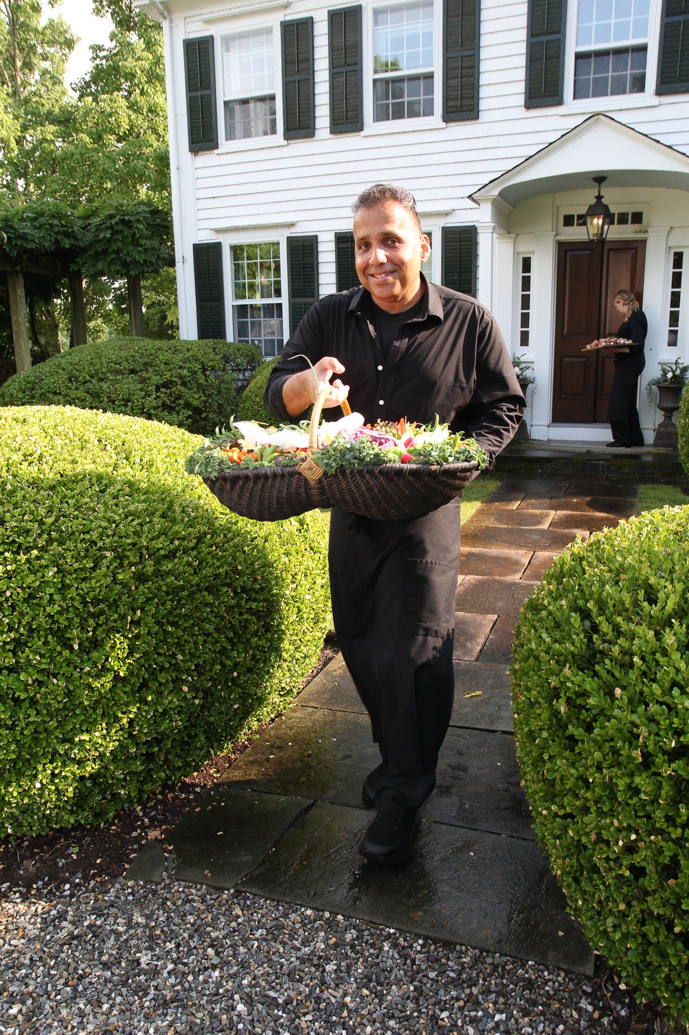 A man in black clothing holding a basket of flowers, smiling, walking on a paved path outside a white house with black shutters, with a woman in black serving food in the background.