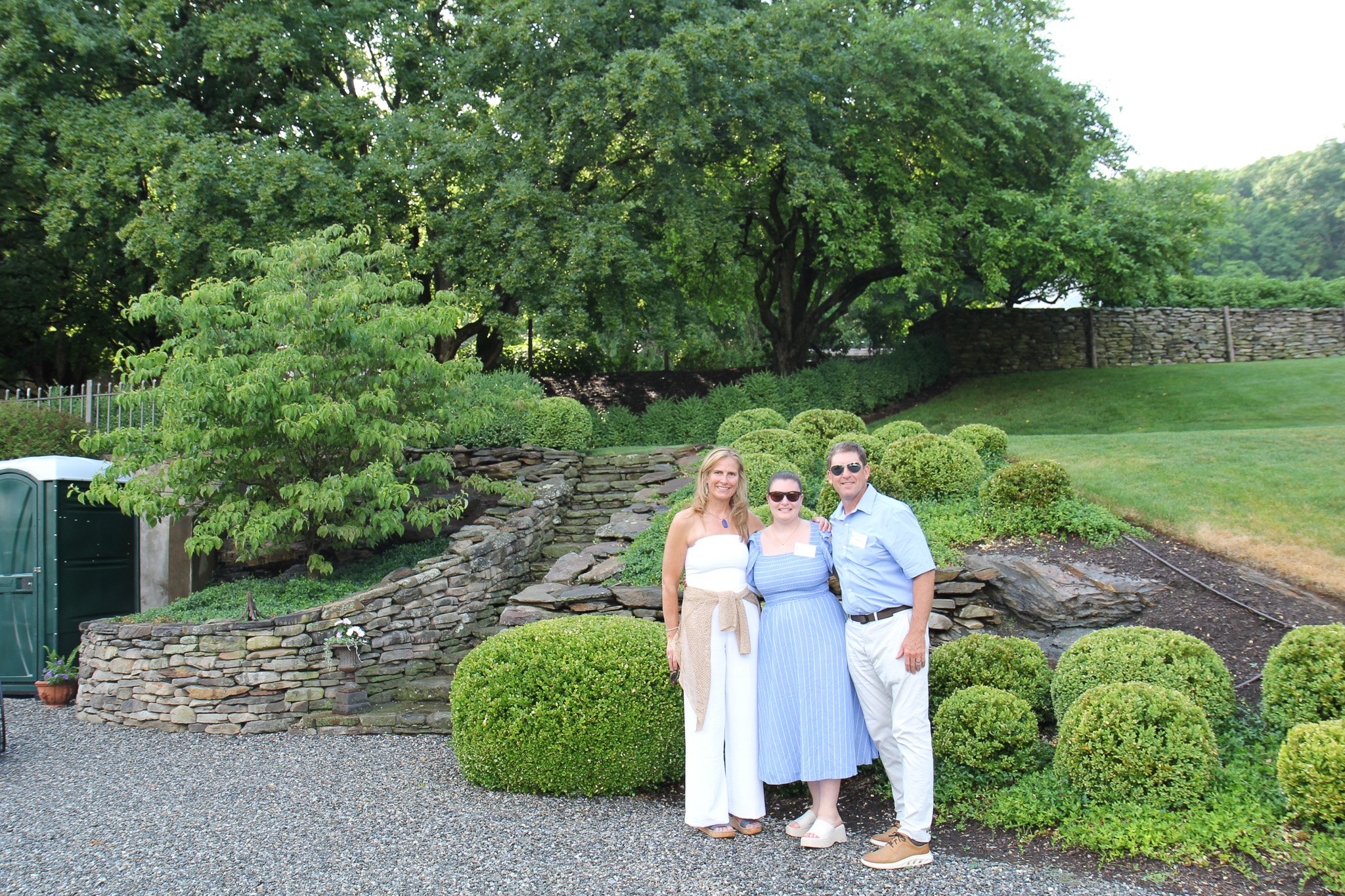 Three people standing outdoors on a gravel path, smiling, with green bushes, large trees, a stone wall, and a grassy area in the background.