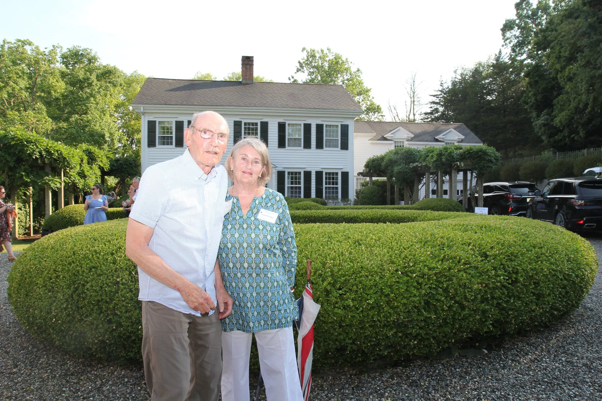 An elderly man and woman standing together outdoors in front of a neatly trimmed hedge, with a white house and parked cars behind them, during daylight.