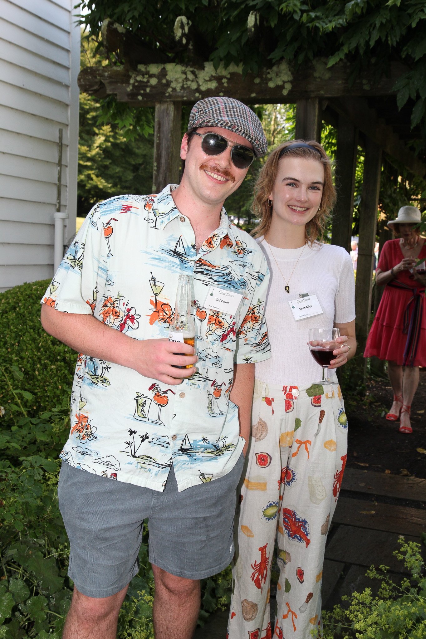 A young man and woman stand outdoors at a social gathering, smiling and holding drinks. The man wears sunglasses, a patterned short-sleeve shirt, gray shorts, and a checkered cap. The woman wears a white top and wide-leg pants with a colorful print. 