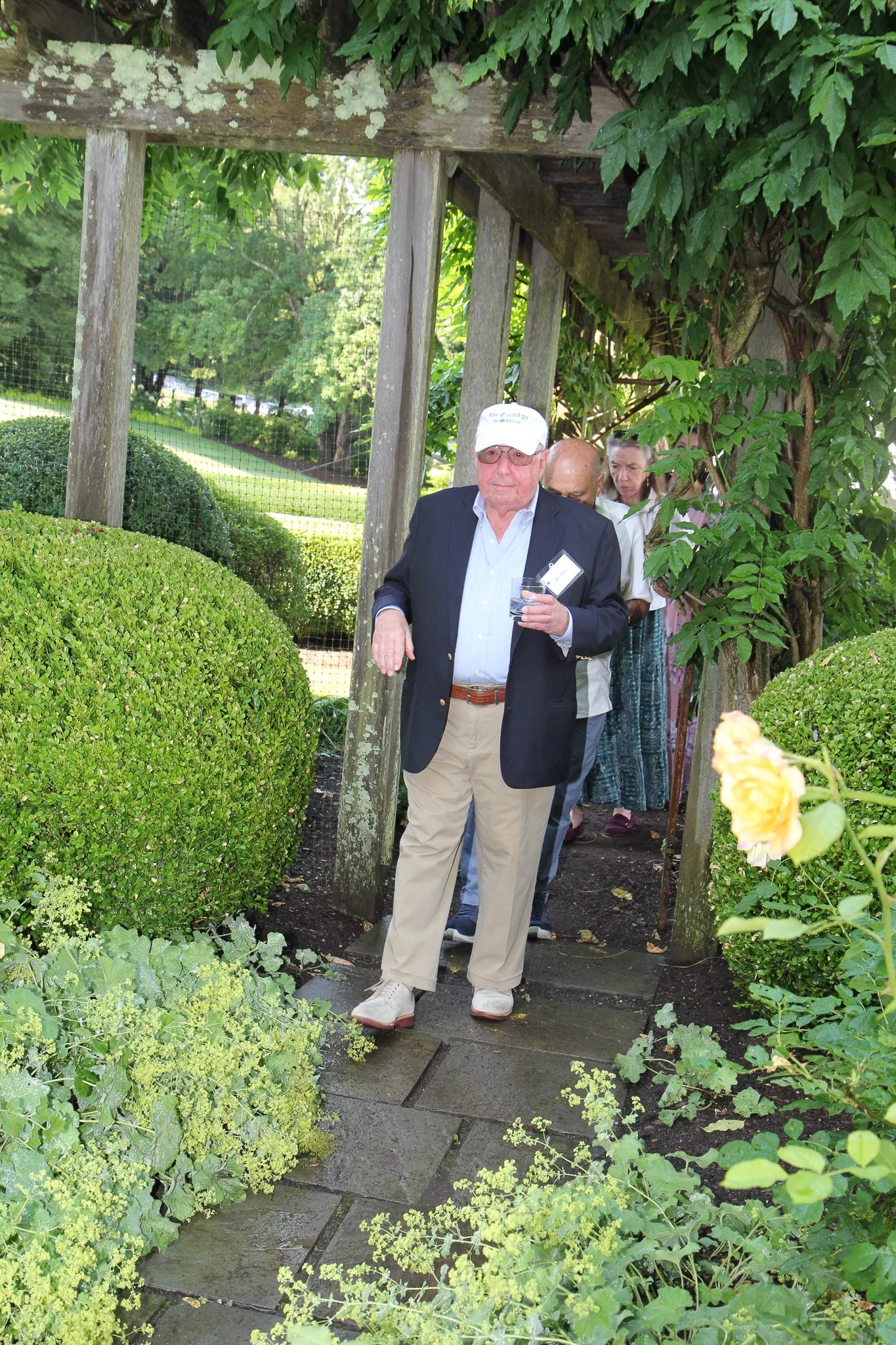 An elderly man in a dark blazer, khaki pants, and a white cap stands on a stone pathway outdoors. He is holding a glass of water and has a name tag. Behind him, a line of people is walking through a garden archway surrounded by lush green plants and 