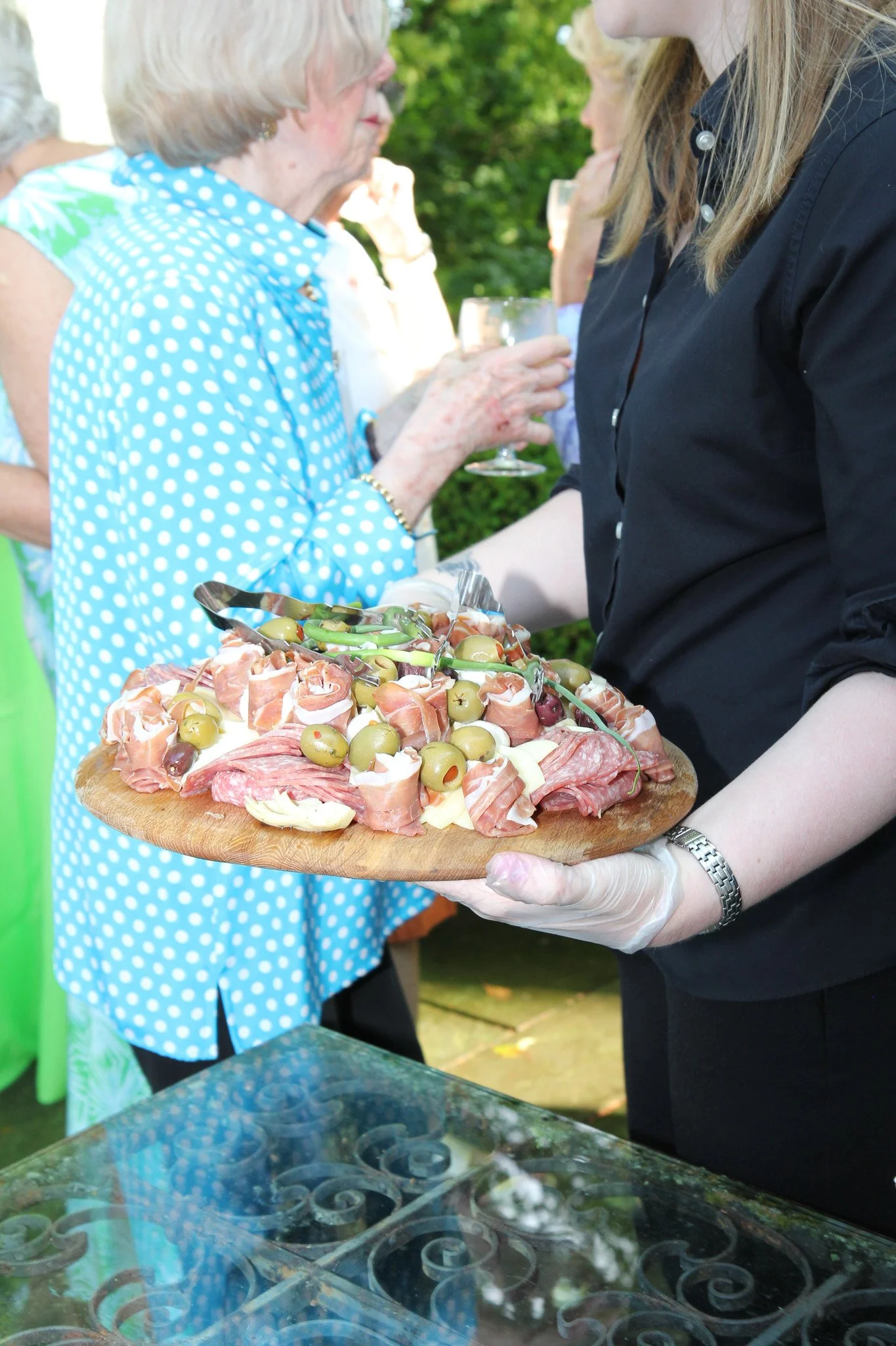 A server in black holding a wooden platter of cured meats, green olives, and cheese slices at an outdoor gathering, with guests in the background holding drinks.