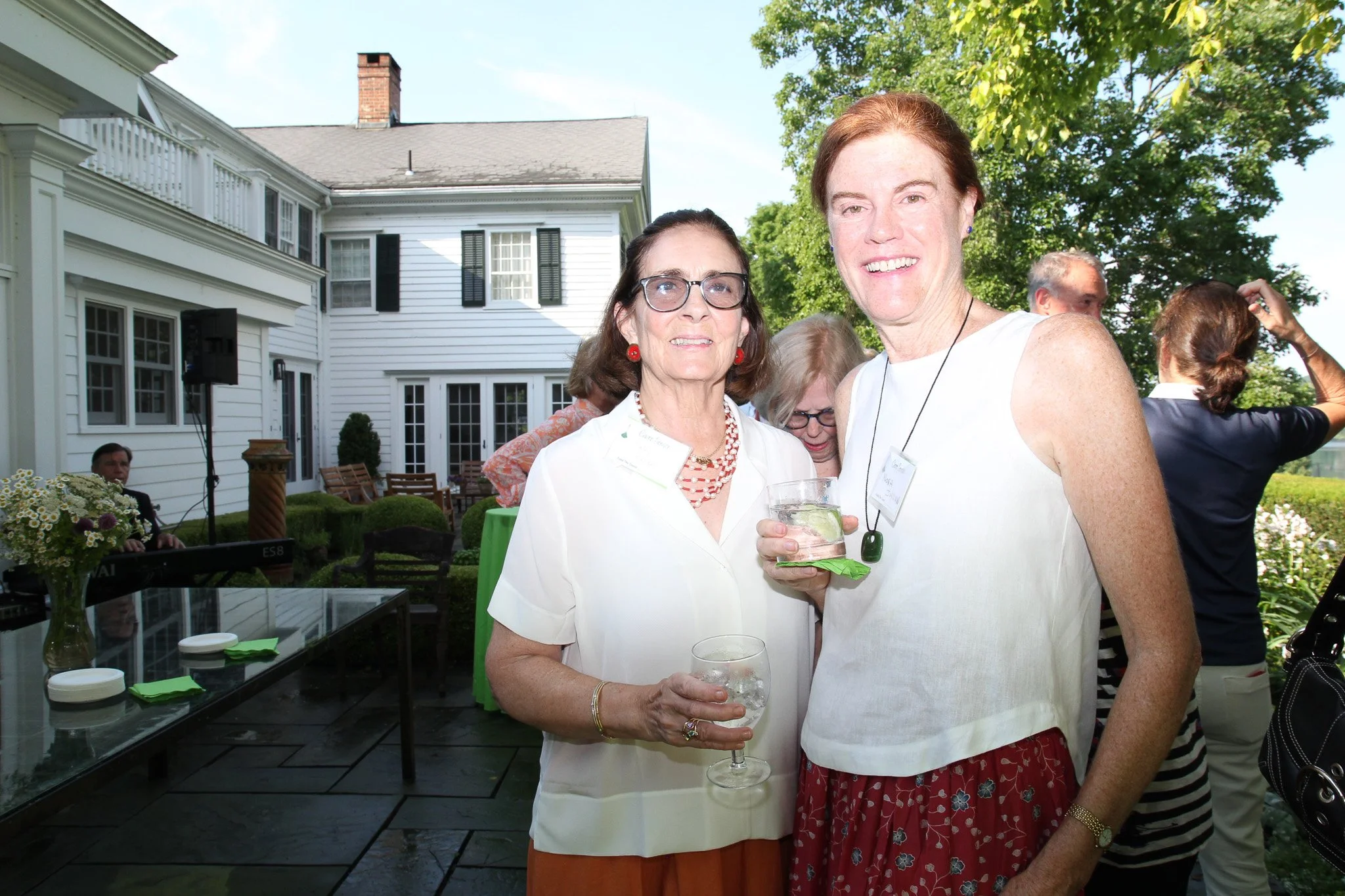 Two women standing outdoors at a social gathering, holding drinks, with a white house and lush greenery in the background.