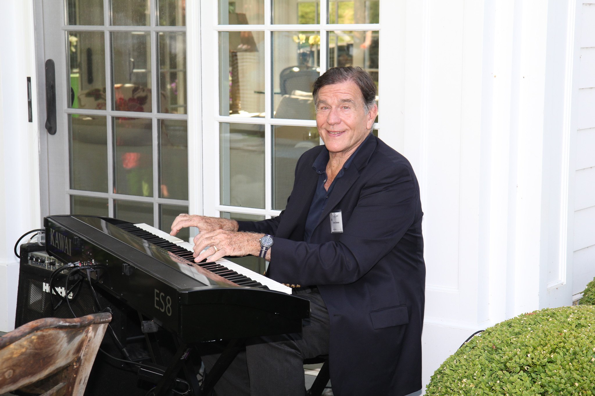 A man wearing a dark suit and a watch, playing a black Kawai keyboard outdoors in Litchfield County near a white building with glass doors.
