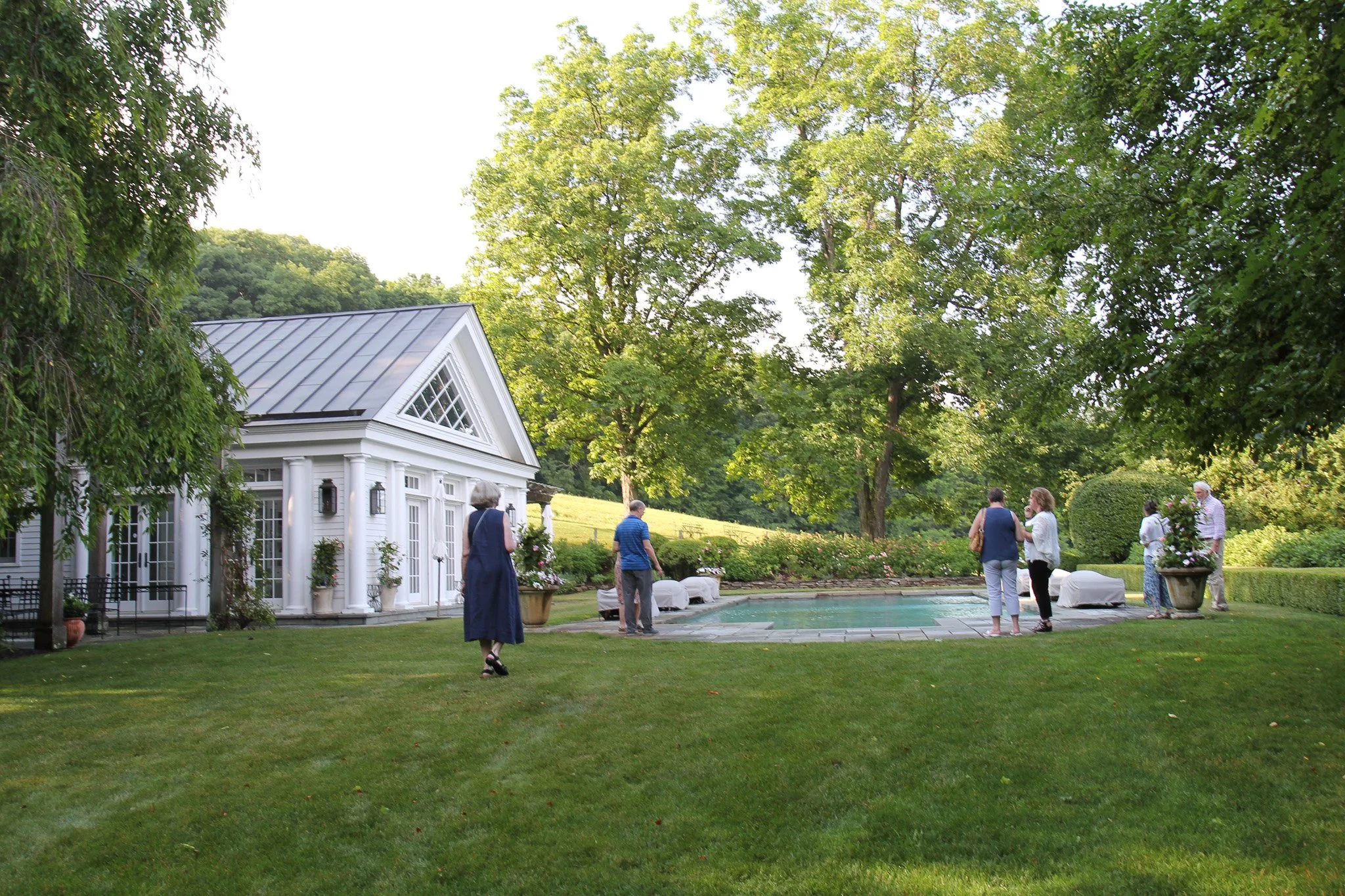 People gather around a swimming pool in a lush, green backyard in Litchfield County with a white house, trees, and shrubs.