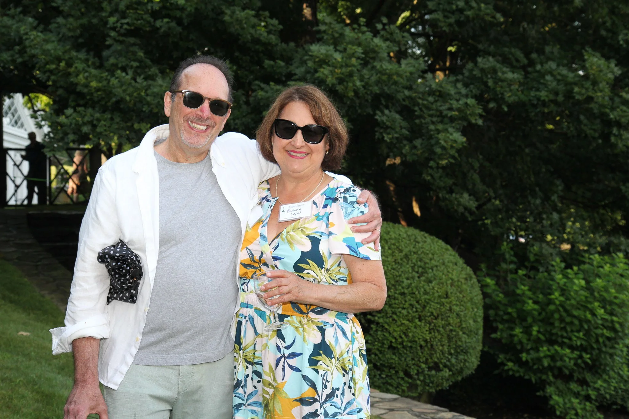 A man and woman standing outdoors in Litchfield County, smiling, with greenery in the background. The woman is holding a glass of wine.