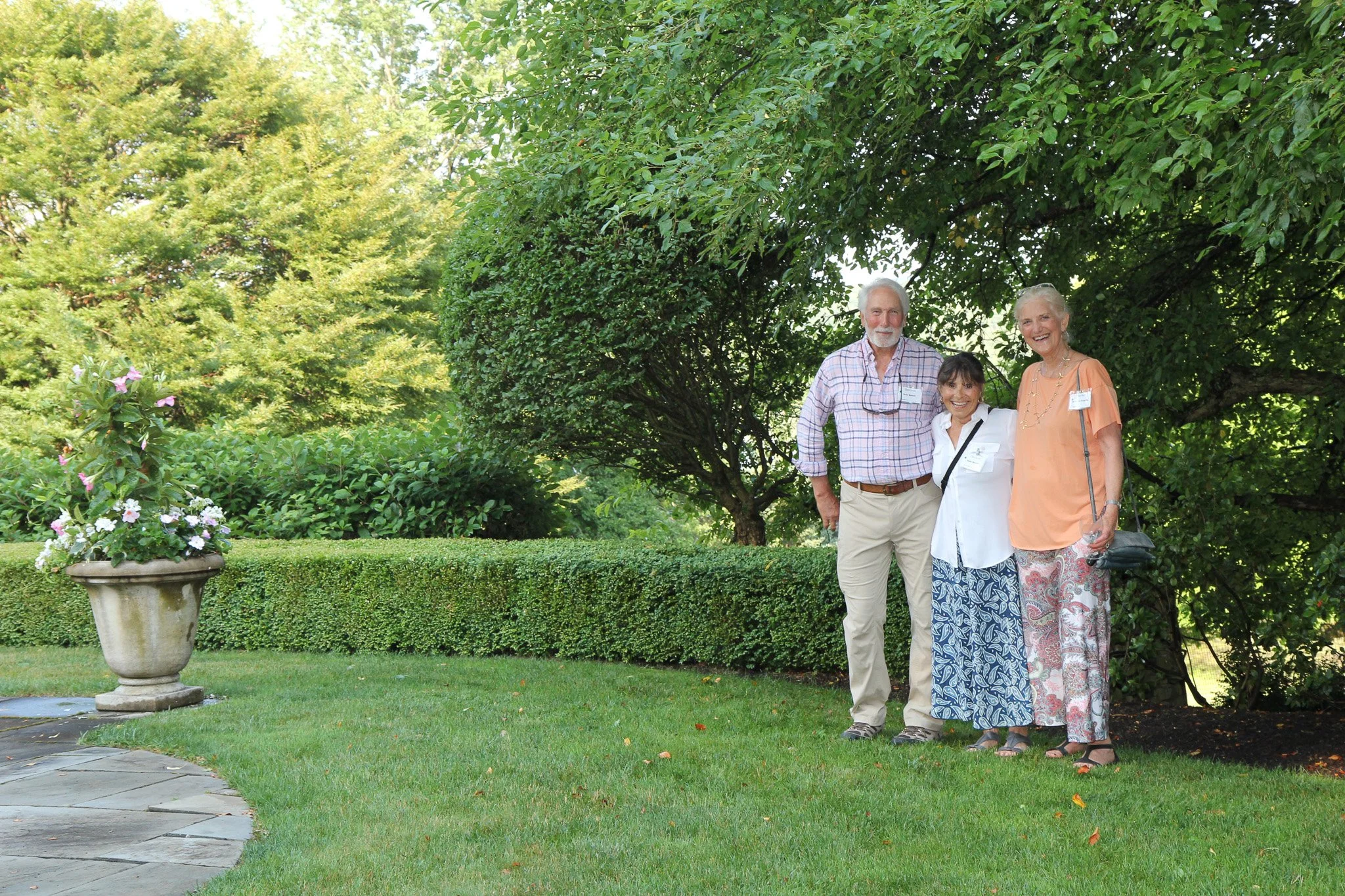 Three adults in Litchfield County standing together outdoors in a garden with a large tree and greenery in the background, smiling at the camera.