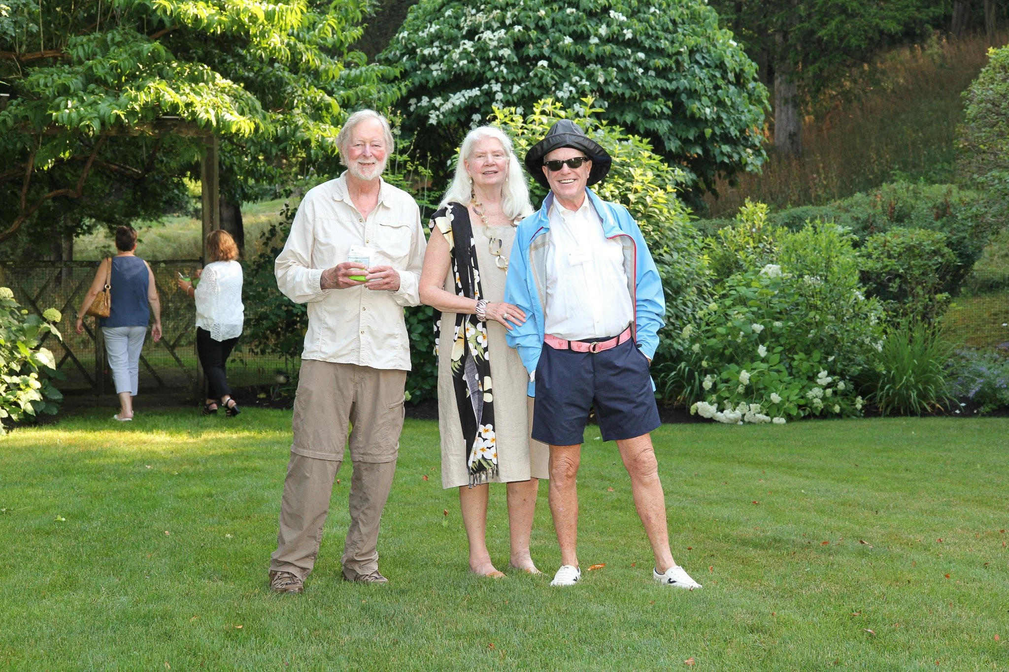 Three seniors standing on a lawn, smiling at the camera, with two additional people in the background near a fence and greenery.