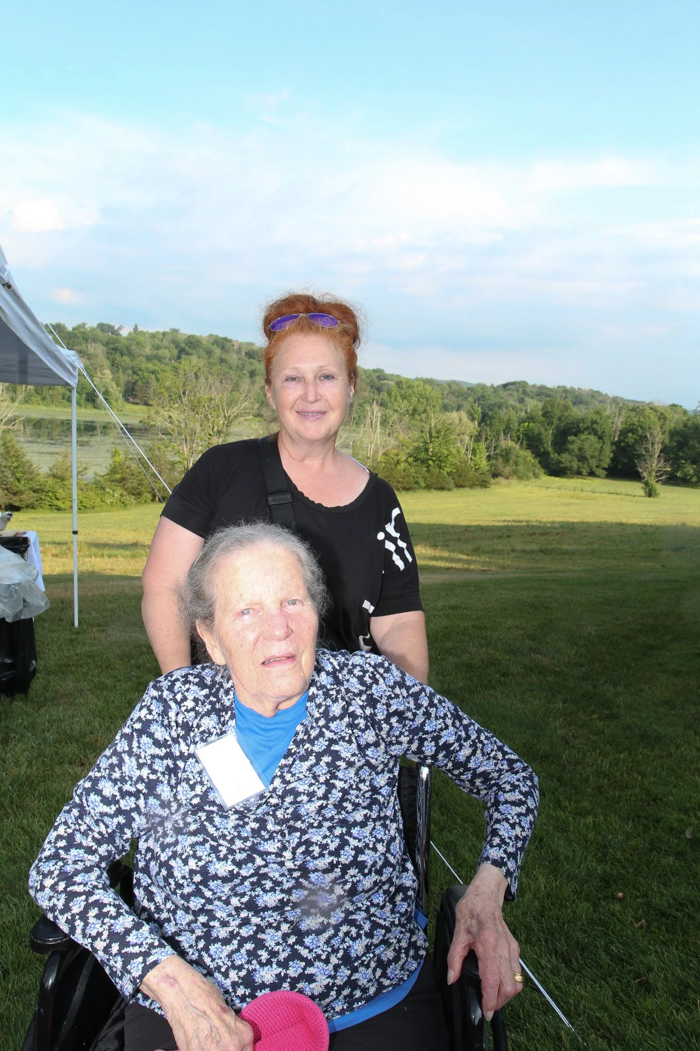 A young woman with red hair and sunglasses on her head standing behind an elderly woman in a wheelchair outdoors in a green field with trees and blue sky in the background.