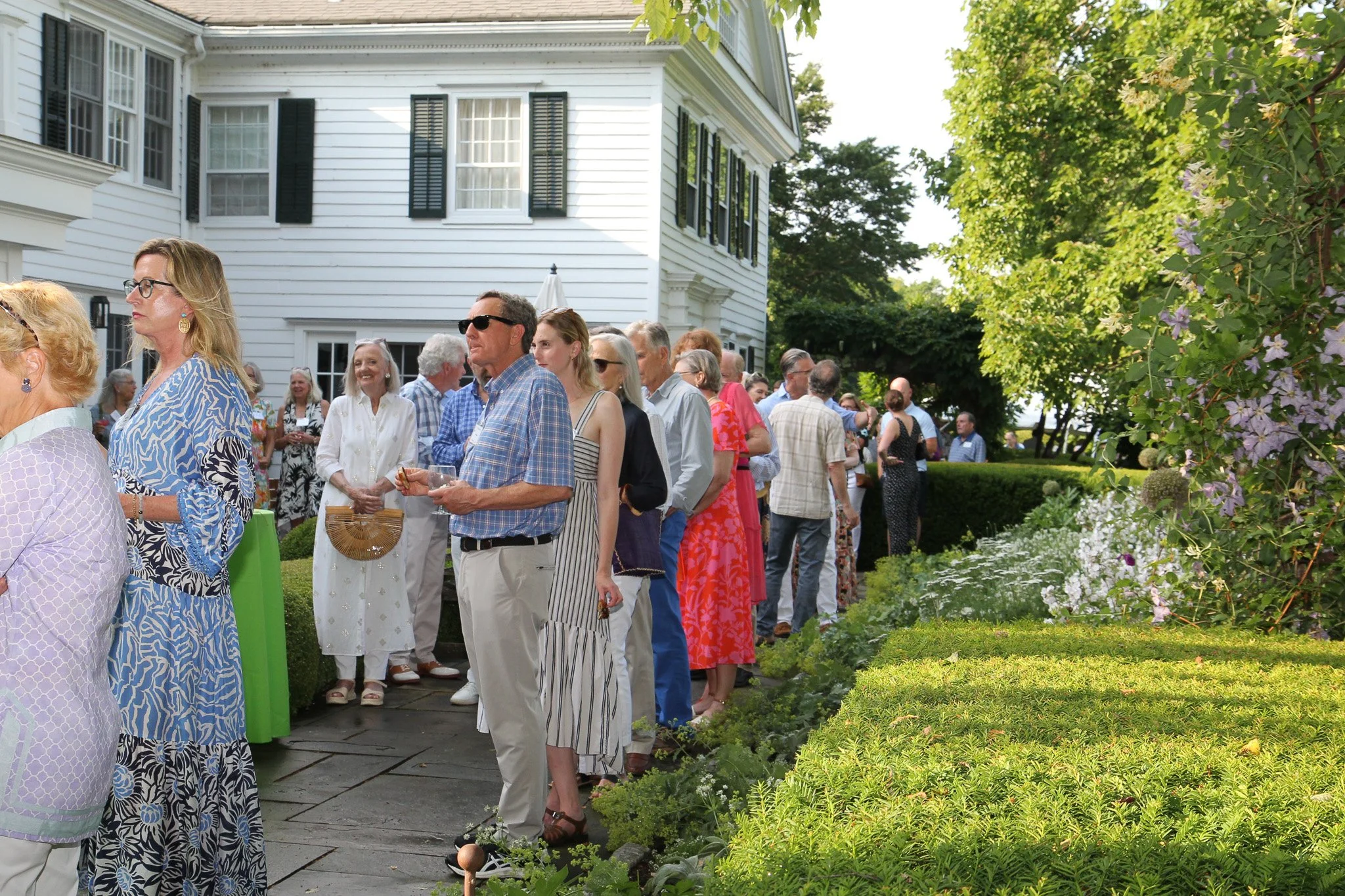 People gathered outdoors at a garden party on a sunny day, standing in line near a white house with black shutters and greenery.