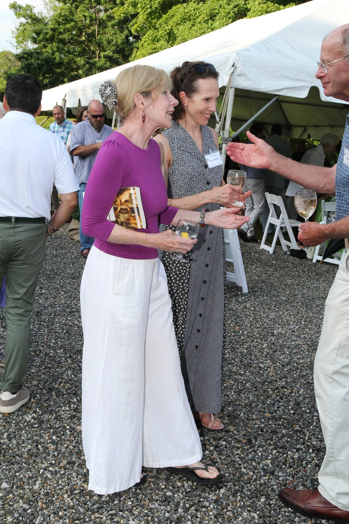 People socializing at an outdoor event under a white canopy tent, engaging in conversations with drinks in hand.