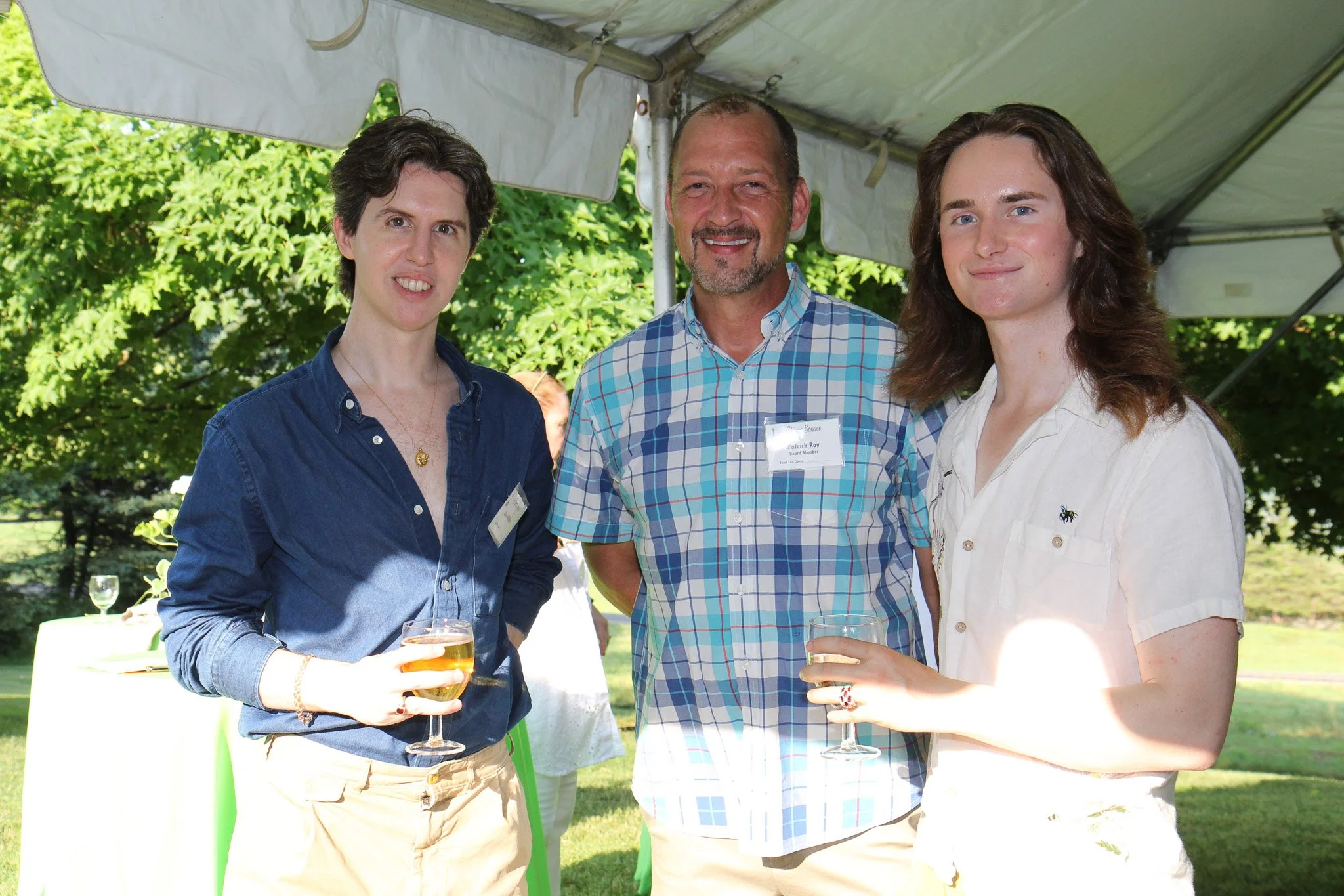 Three people standing under a tent outdoors, holding glasses of white wine, smiling at the camera, with greenery in the background.