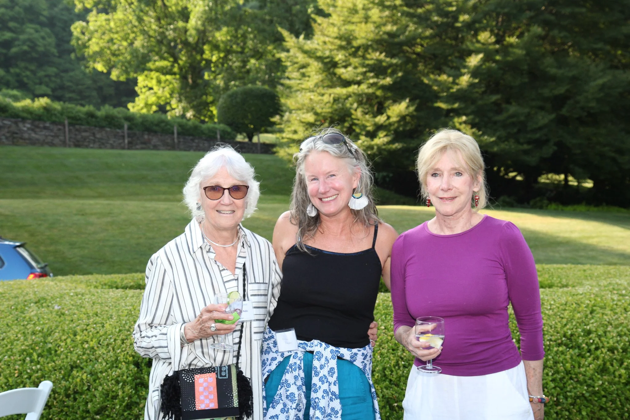 Three women standing outdoors in a garden, smiling and holding drinks, with trees and greenery in the background.