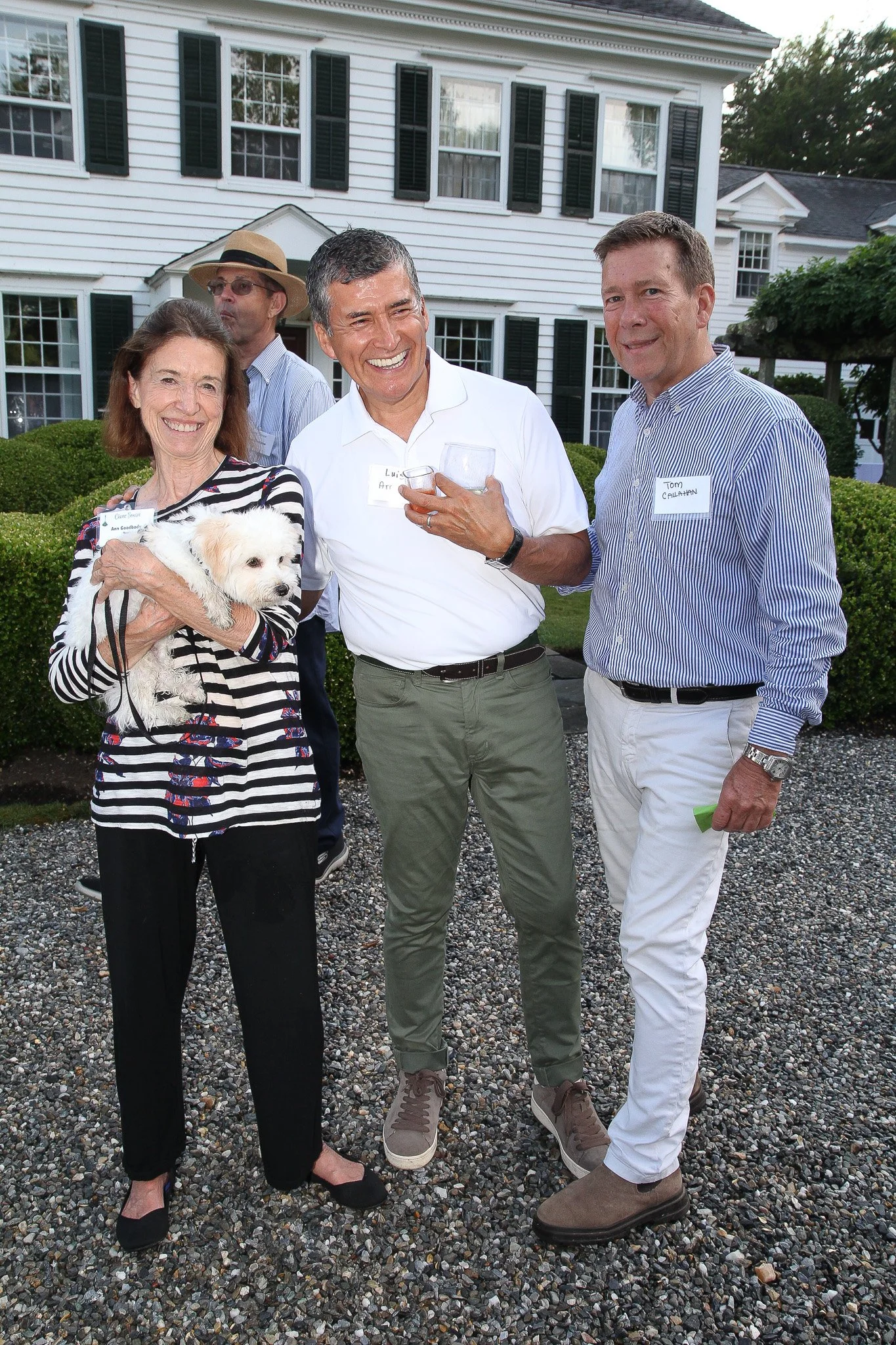 Group of four people and a dog at an outdoor gathering in front of a large white house with black shutters. The woman on the left is holding a small white dog. The three men are smiling, with two of them holding drinks.
