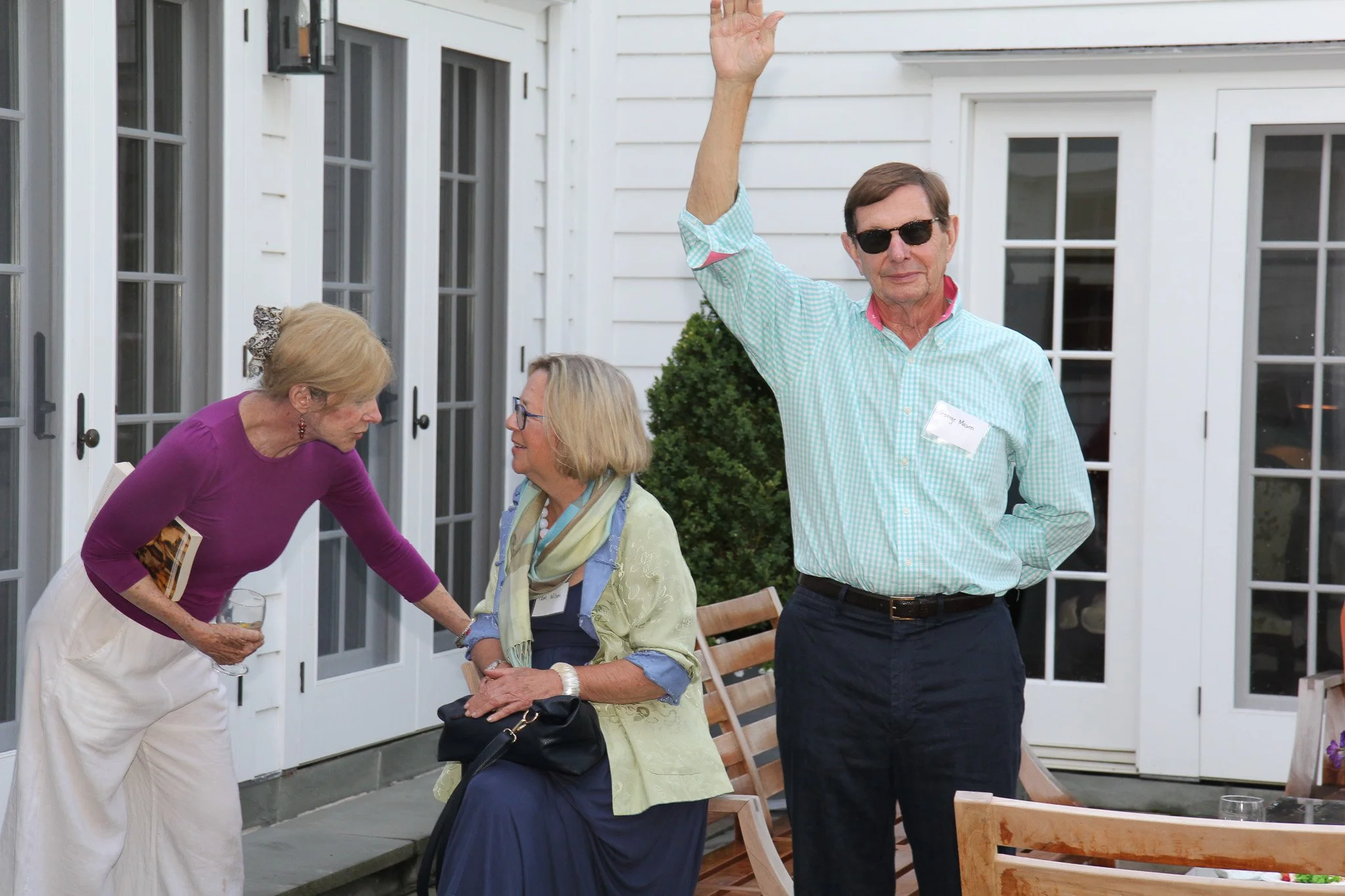 Three older adults at an outdoor gathering, one woman leaning in to speak to a seated woman, and a man standing with his hand raised, all on a patio with white doors and greenery.