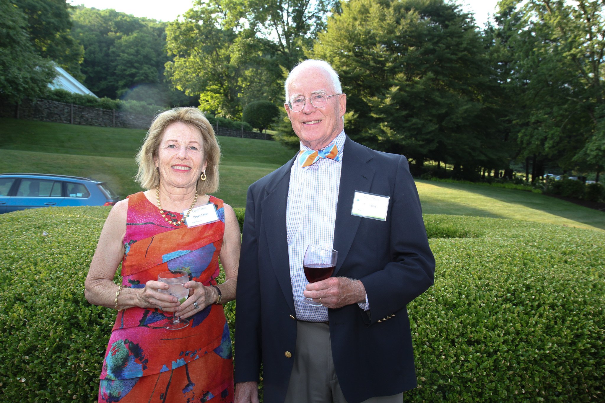 An elderly woman and man smiling outdoors at a social gathering, holding drinks, with greenery and trees in the background.