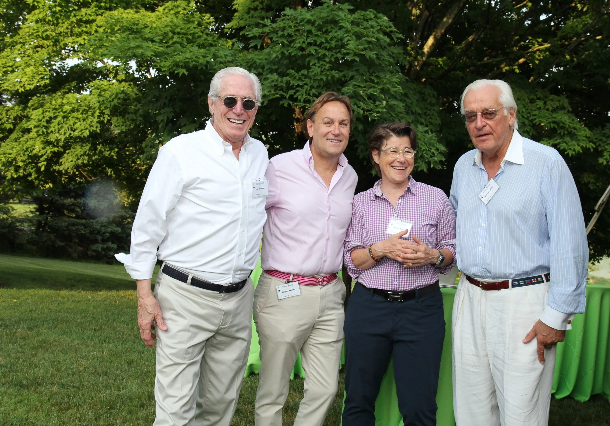 Four smiling adults standing outdoors on a grassy area with trees in the background, wearing casual business attire and name badges.