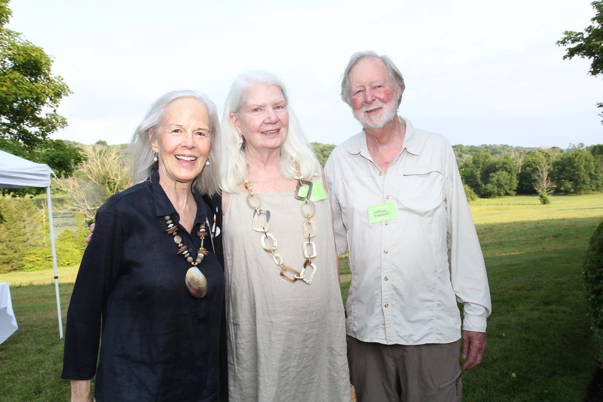 Three elderly adults, two women and one man, standing outdoors on a grassy field with trees in the background, smiling at the camera, dressed casually with name tags, during a daytime gathering or event.
