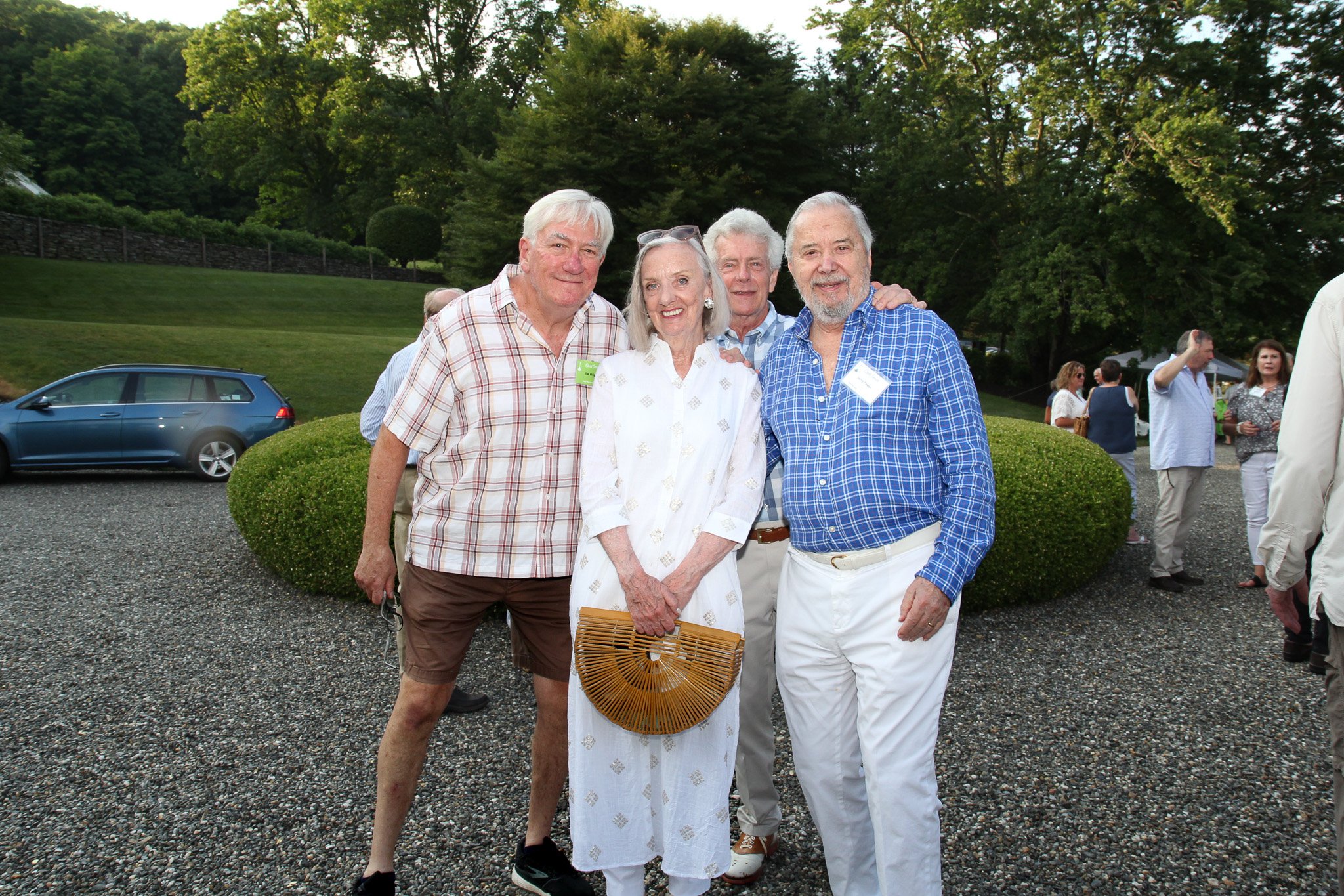 Four elderly adults smiling and posing for a photo outdoors at a social gathering, with greenery and other people in the background.
