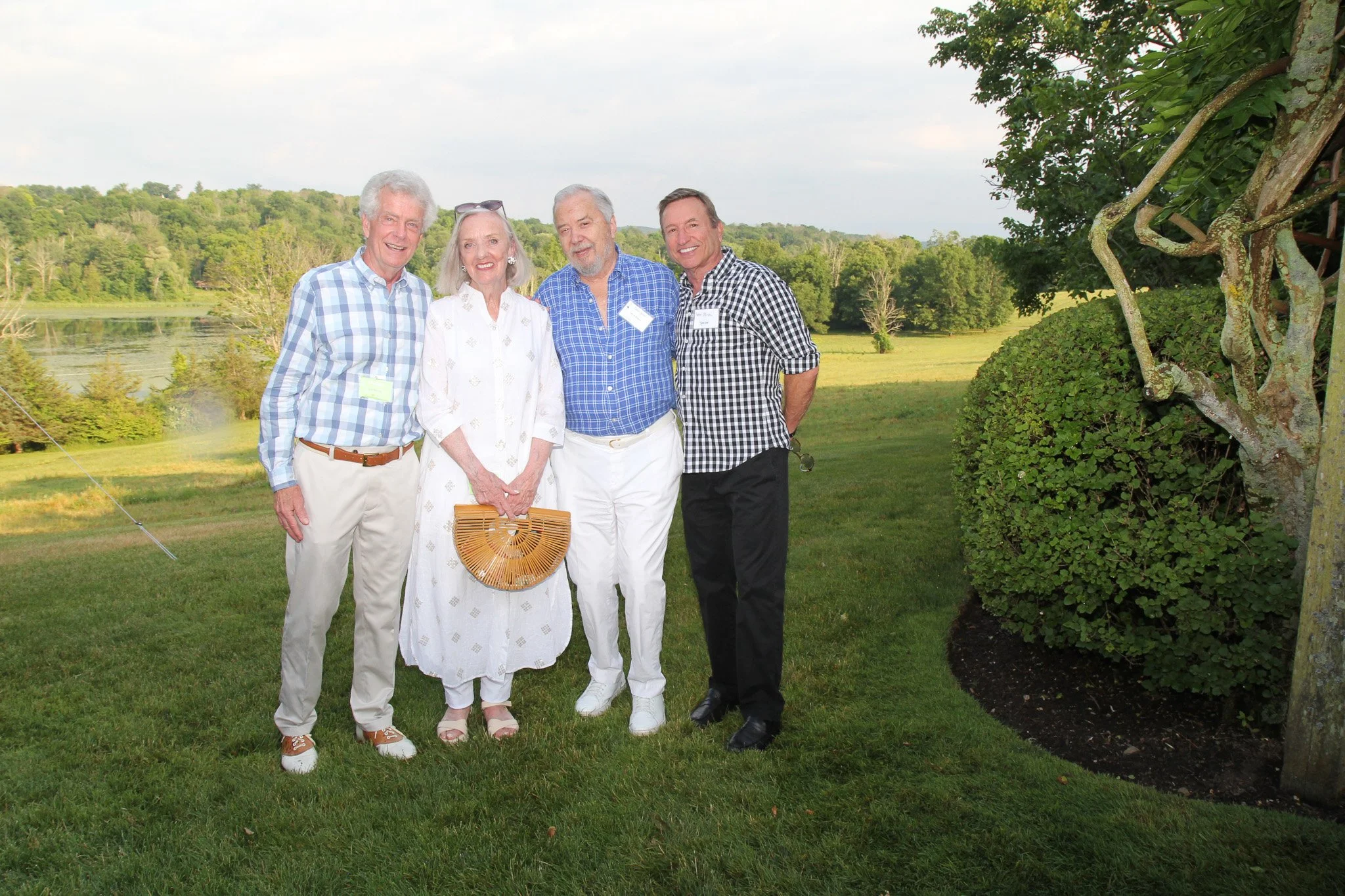 Four people standing outdoors on grass near a tree, with a lake and trees in the background, smiling for the camera.