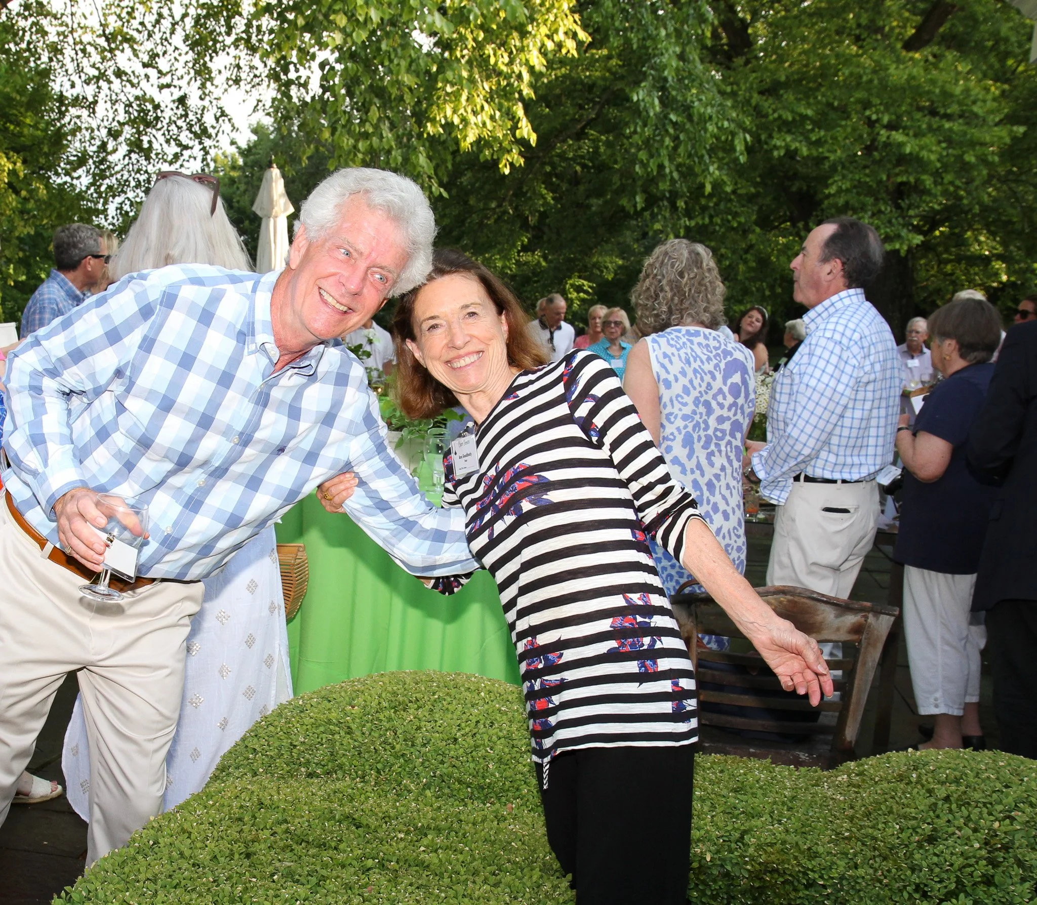 Two smiling elderly people, a man and a woman, at an outdoor social event. The man has white hair and is wearing a light blue checkered shirt and beige pants. The woman has shoulder-length brown hair and is wearing a black and white striped dress wit