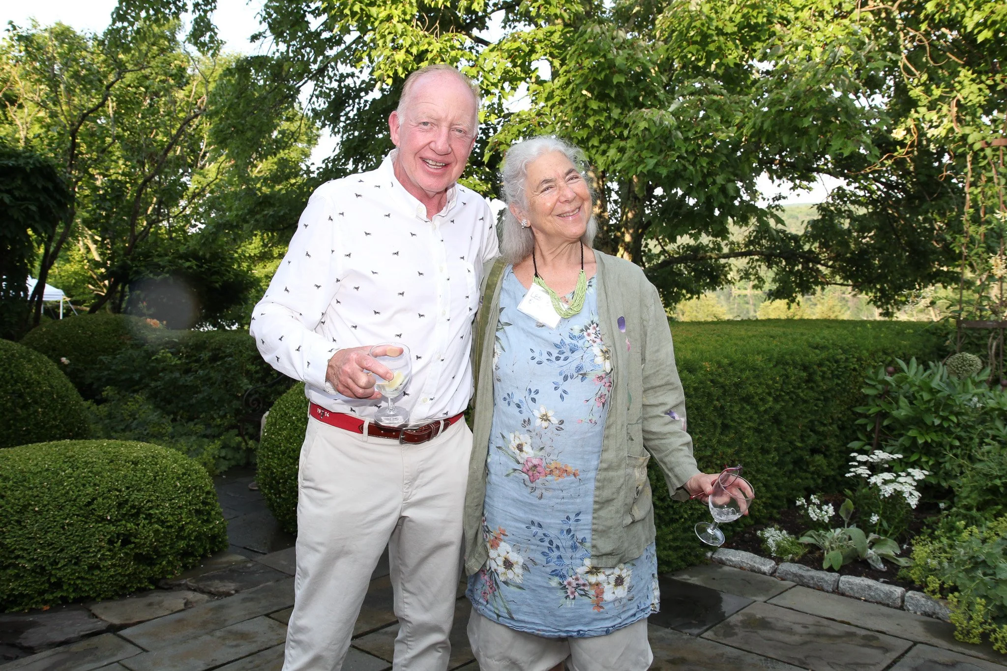 An elderly man and woman standing outdoors in a garden, smiling and holding wine glasses, with trees and bushes in the background.