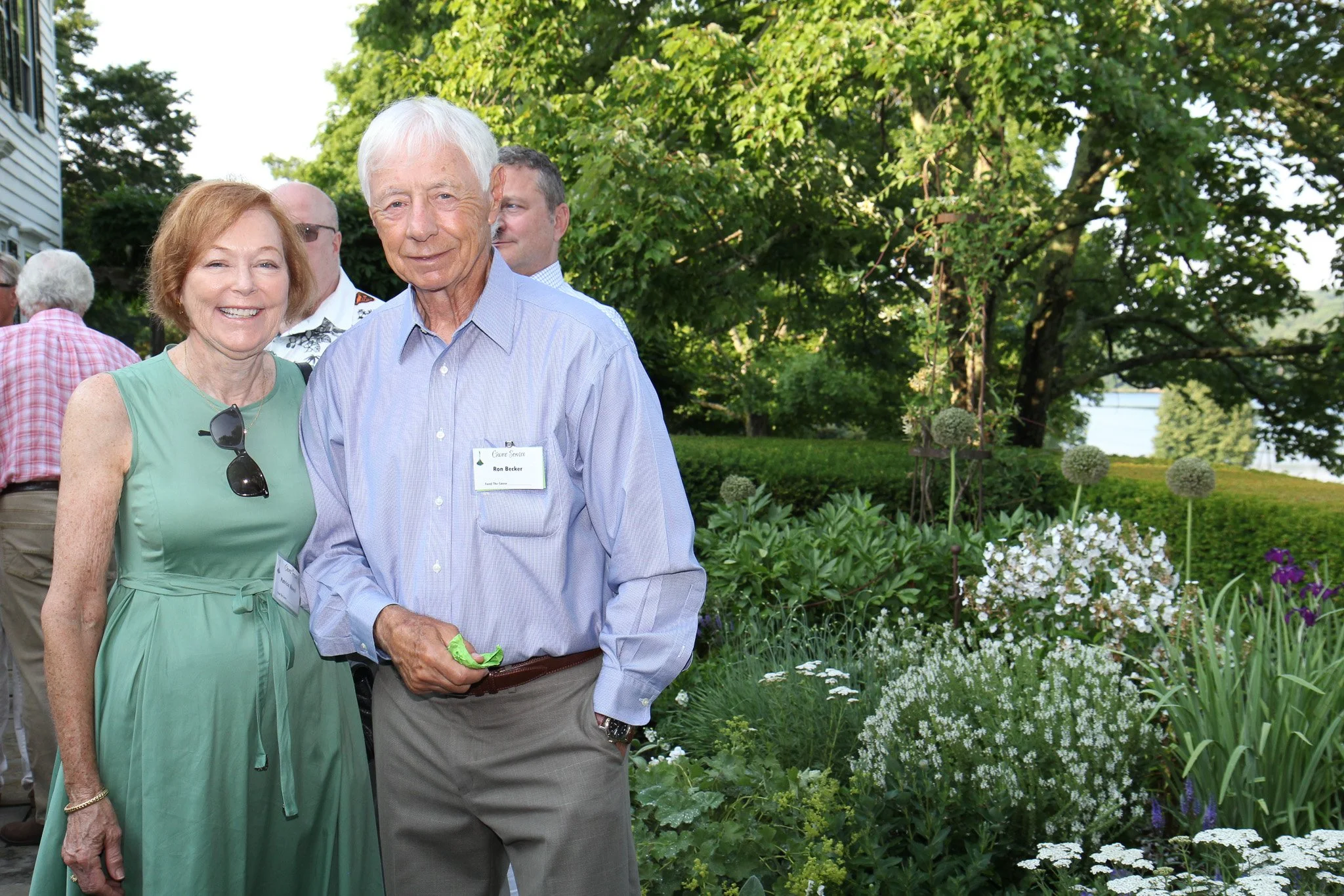 A smiling woman in a green sleeveless dress and sunglasses hanging from her dress, standing next to a man in a light purple dress shirt and beige pants, outdoors with trees and flowers in the background.