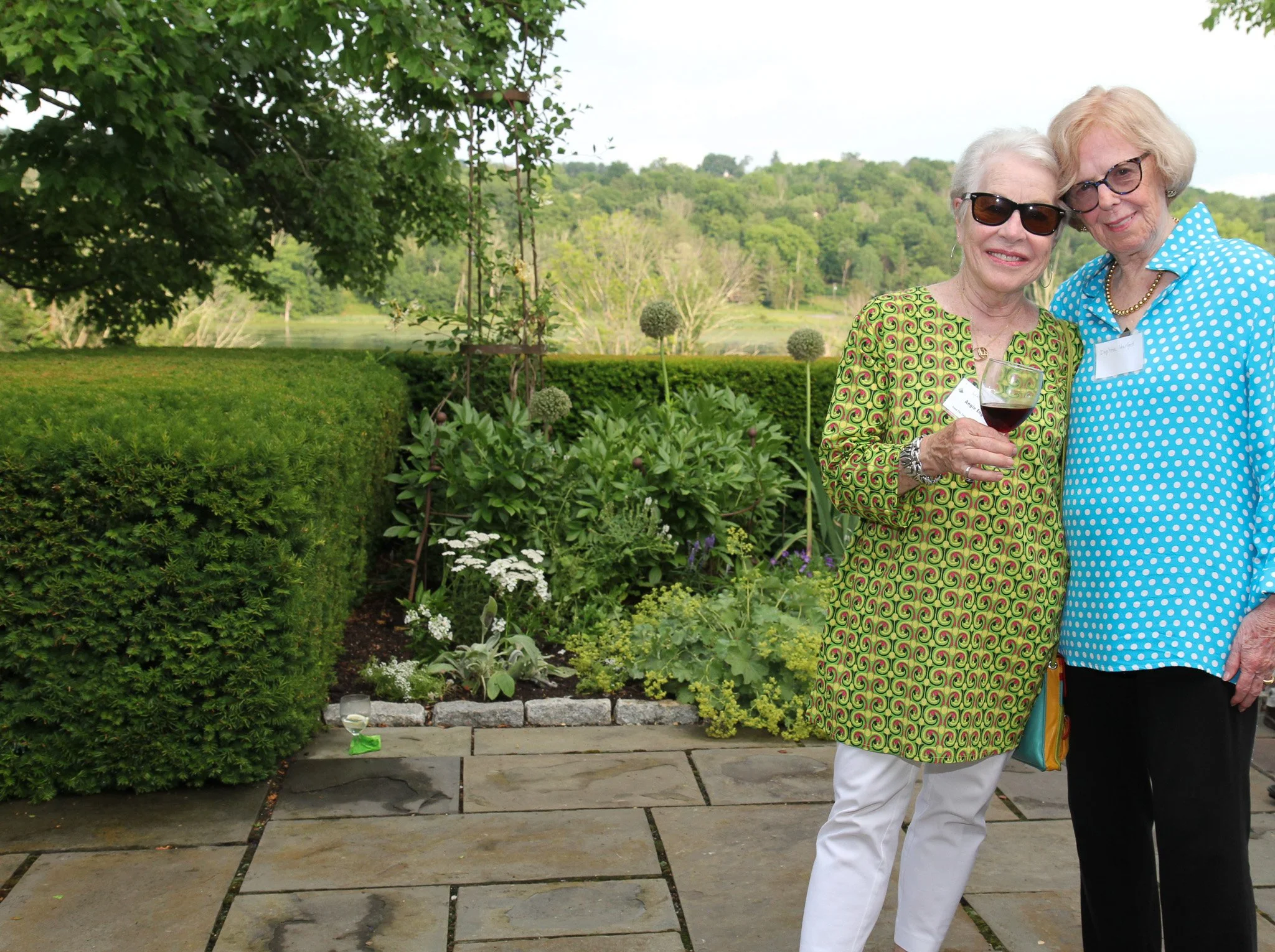 Two elderly women standing together outdoors on a patio, smiling at the camera, one holding a glass of red wine, with a lush garden and scenic landscape in the background.