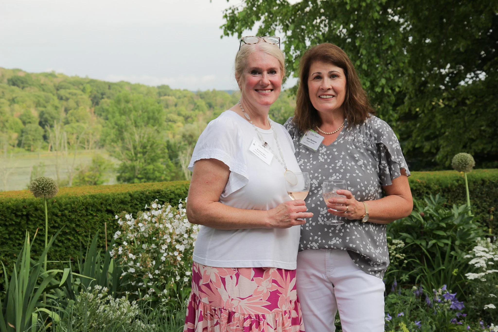 Two women standing outdoors in front of a garden holding wine glasses, smiling at the camera.