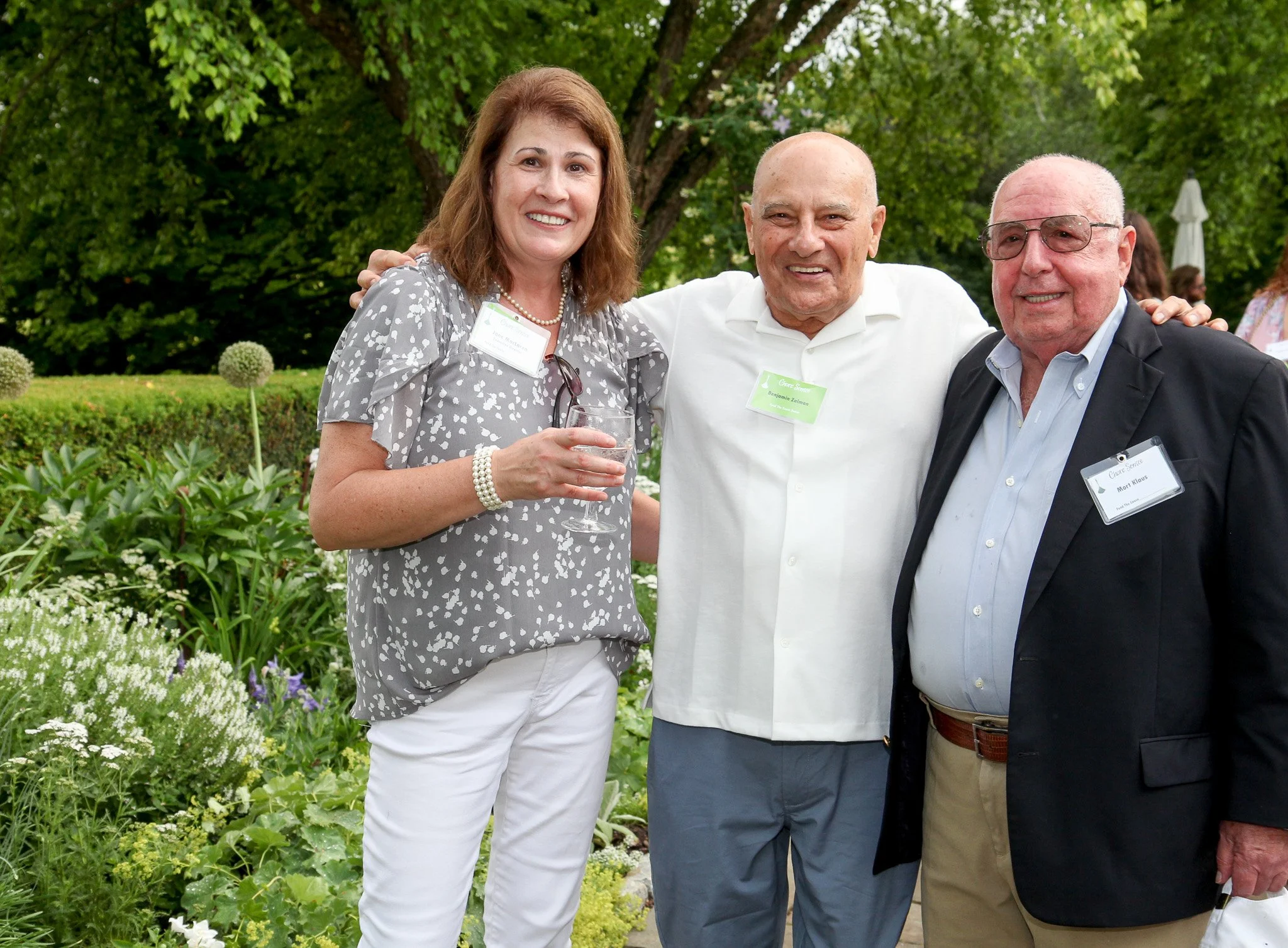Three people posing at an outdoor event, standing in front of lush green trees and plants. The woman on the left is holding a wine glass, wearing a gray patterned blouse, white pants, pearl bracelet, and glasses hanging from her collar. The man in th