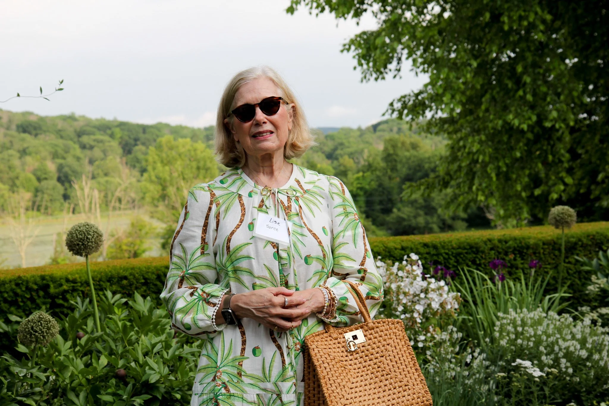 An elderly woman with blonde hair, wearing dark sunglasses and a patterned blouse with green leaves and animals, stands outdoors in a garden with lush greenery and white and purple flowers. She has a tan woven handbag and wears jewelry, including bra