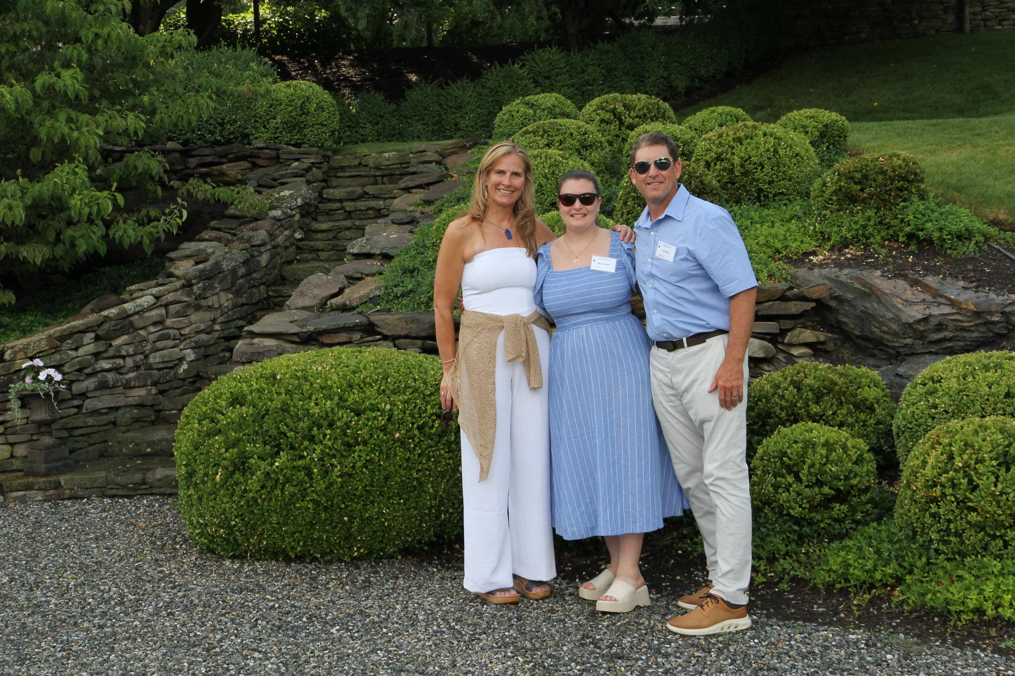 Three people standing outdoors on a gravel path, smiling, with green bushes and a rock wall in the background.
