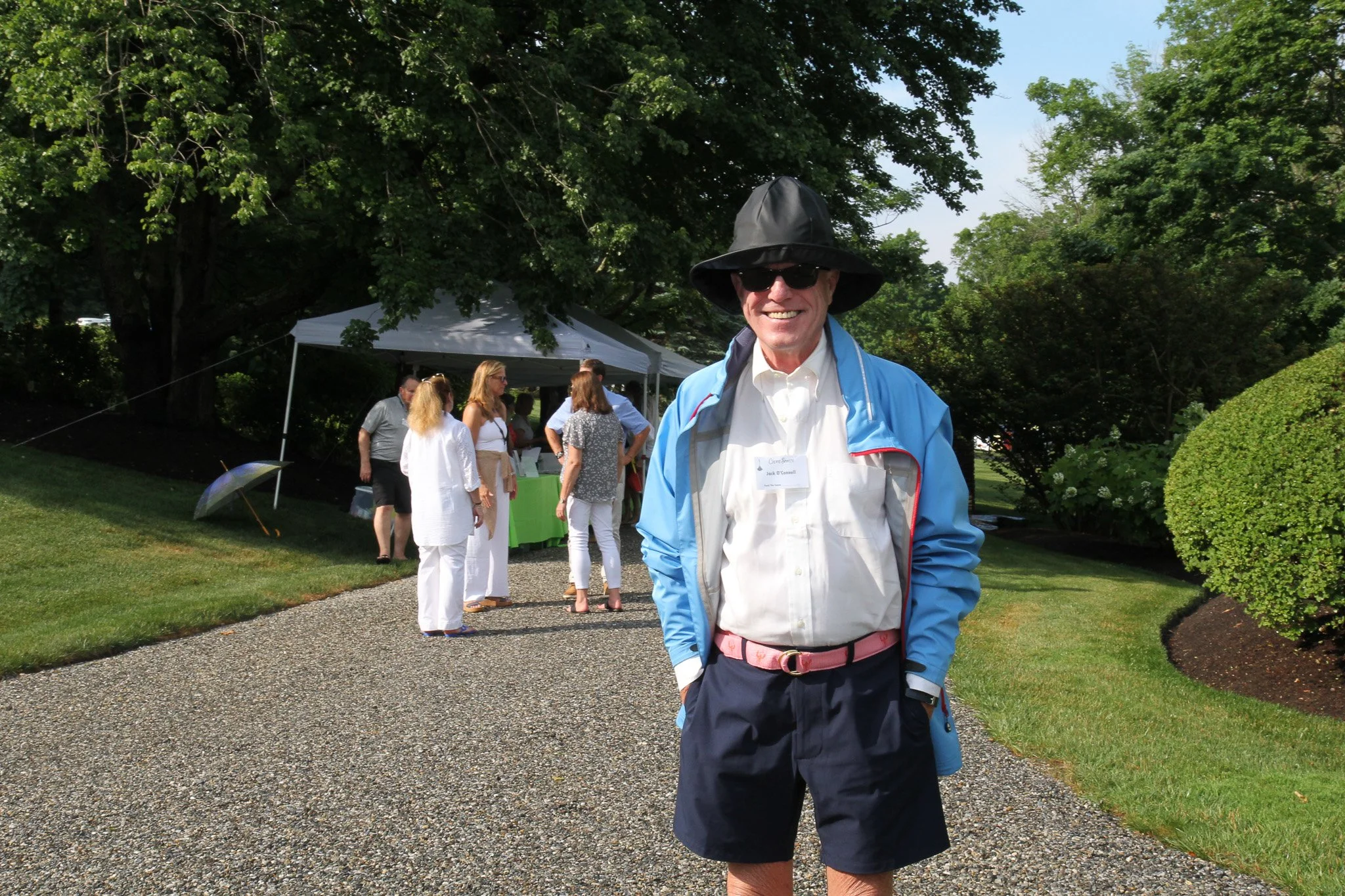 Smiling man wearing sunglasses, a black hat, a light blue jacket, with his hands in his pockets at an outdoor gathering, with a group of people under a white canopy in the background