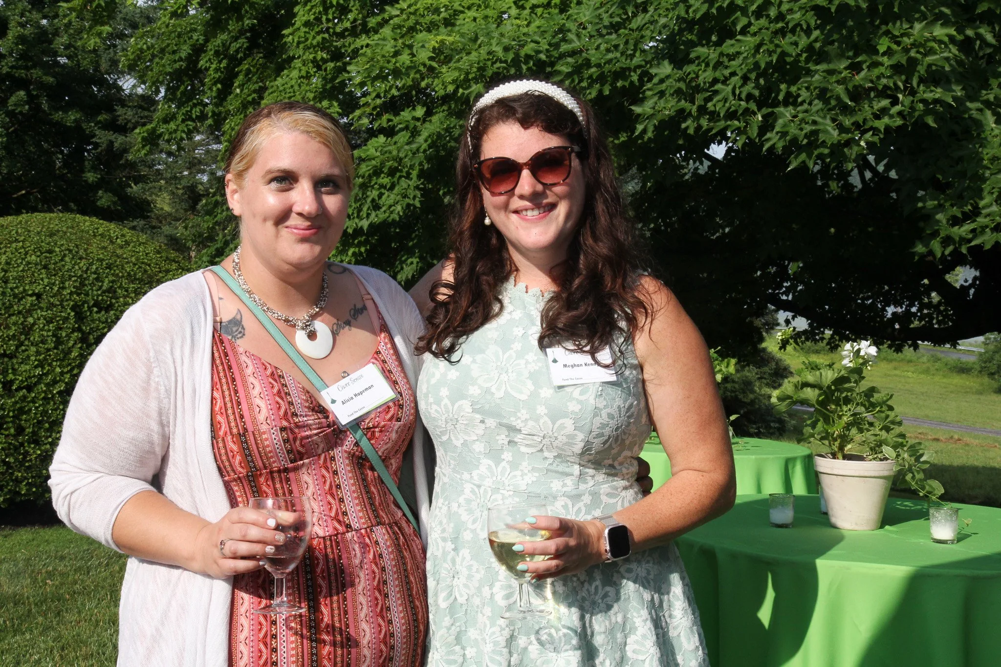 Two women standing outdoors under a large tree, smiling and holding wine glasses, one wearing sunglasses and a white patterned dress, the other wearing a colorful dress with a white cardigan.