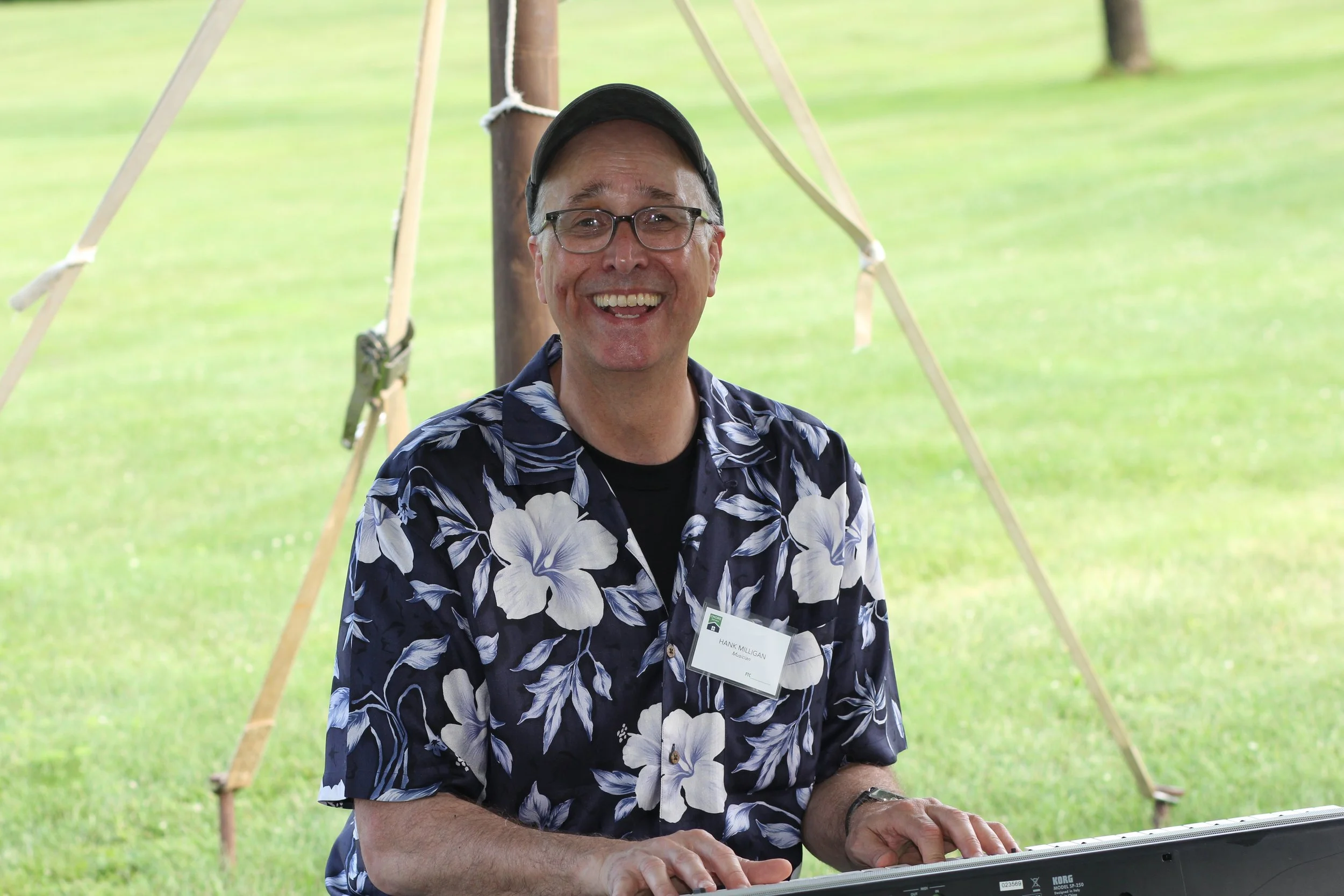 A smiling man wearing glasses, a black cap, and a Hawaiian shirt playing a keyboard outdoors with a wooden post and ropes in the background, on a grassy field.